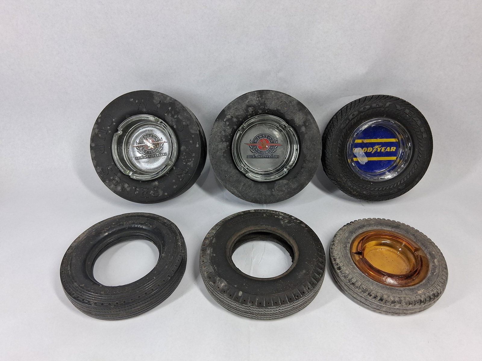 6 Vtg Goodyear & Winston Tire Ashtrays: A collection of six vintage automotive and racing themed collectible tire ashtrays and tire frames. The group features two large black rubber tires with embossed sidewall text reading WINSTON CUP