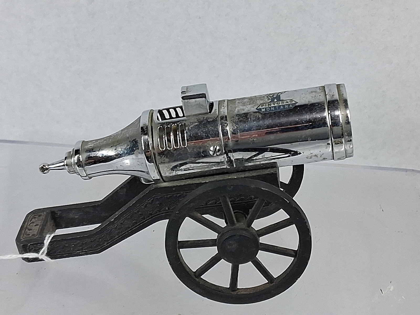 Pinturas Montana Cannon Table Lighter: A vintage promotional table lighter designed in the form of a field cannon. The object features a chrome-plated metal barrel assembly mounted on a dark-finished cast metal carriage with two spoked