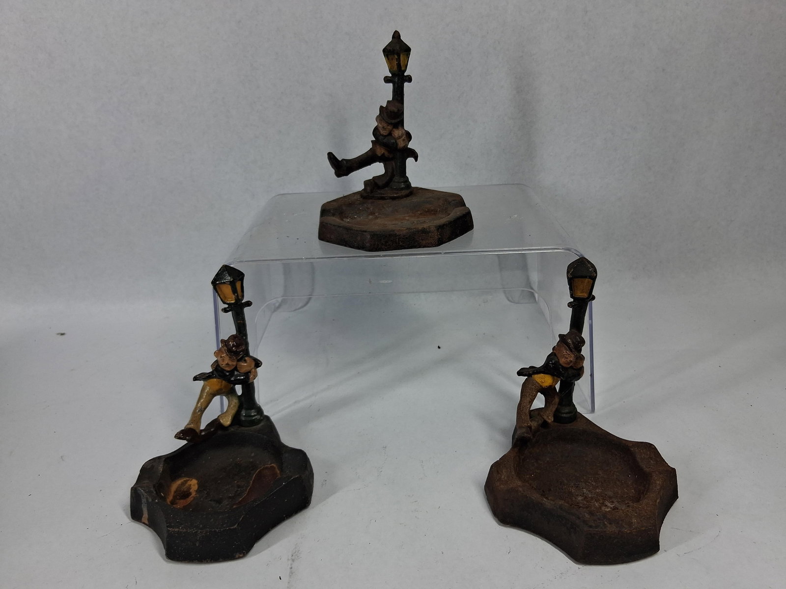 Set 3 Japanese Cast Iron Drunk Ashtrays: A trio of Japanese cast iron novelty ashtrays featuring hand-painted figures. Each ashtray depicts a man wearing a suit and hat leaning against a street lamppost, with one figure shown with a leg