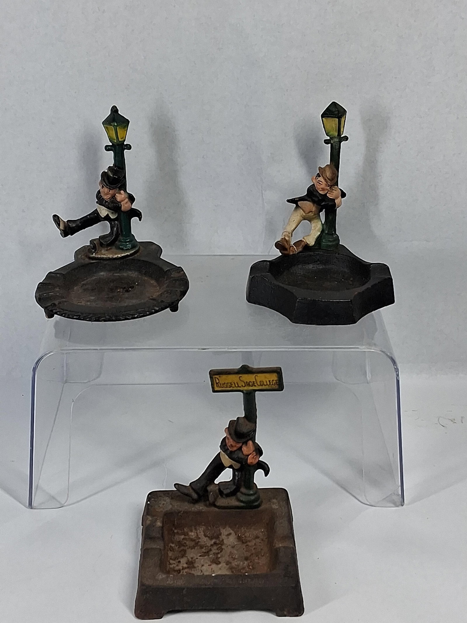 3 Wilton Iron Drunk & Signpost Ashtrays: A trio of vintage cold-painted cast iron novelty ashtrays produced by Wilton Products. Each piece features a figural character in a state of intoxication leaning against a vertical support. The