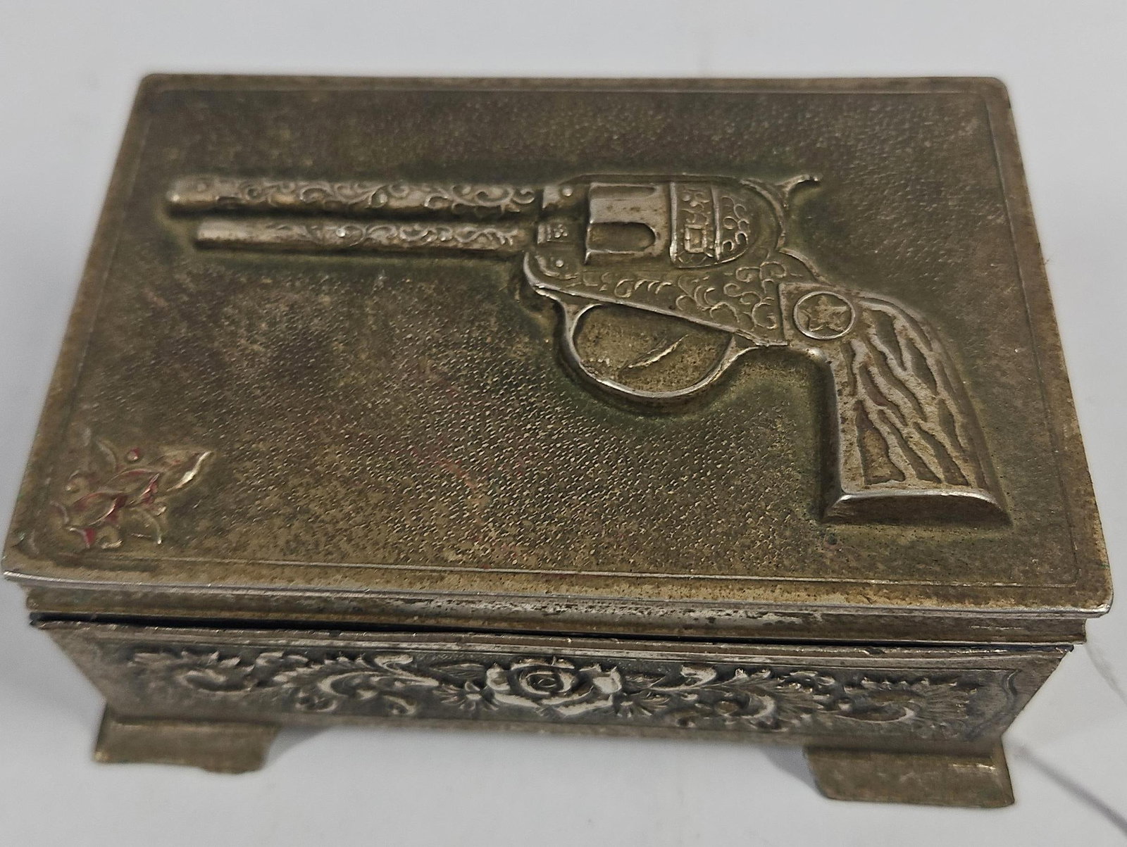 Silver-Plated Western Revolver Box: A rectangular silver-plated metal trinket box featuring a Western design. The hinged lid is ornamented with a high-relief depiction of a single-action revolver decorated with scrollwork and a five-poi