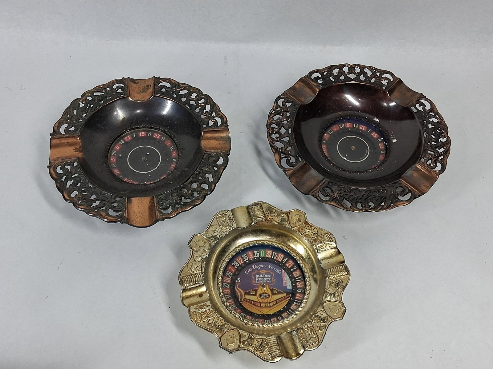 3 Japanese Mechanical Roulette Ashtrays: A trio of Japanese mechanical souvenir ashtrays featuring integrated roulette wheels. Two matching pieces are constructed with copper-toned openwork filigree rims and black enameled bowls. Each filigr