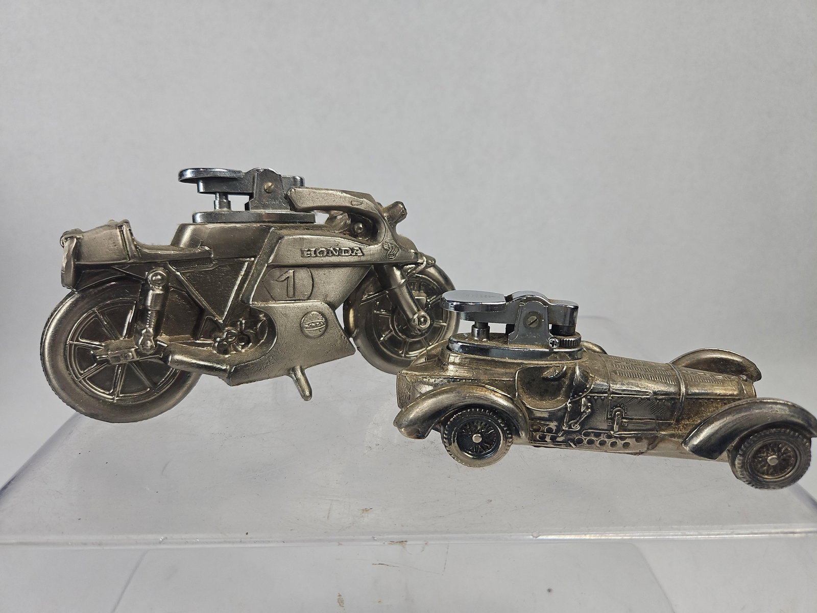 Pair Japanese Honda & Bugatti Lighters: A lot of two vintage Japanese cast metal tabletop cigarette lighters with a silver-tone finish. The first item is a racing motorcycle featuring a full fairing, spoked wheel detailing, and an integrate