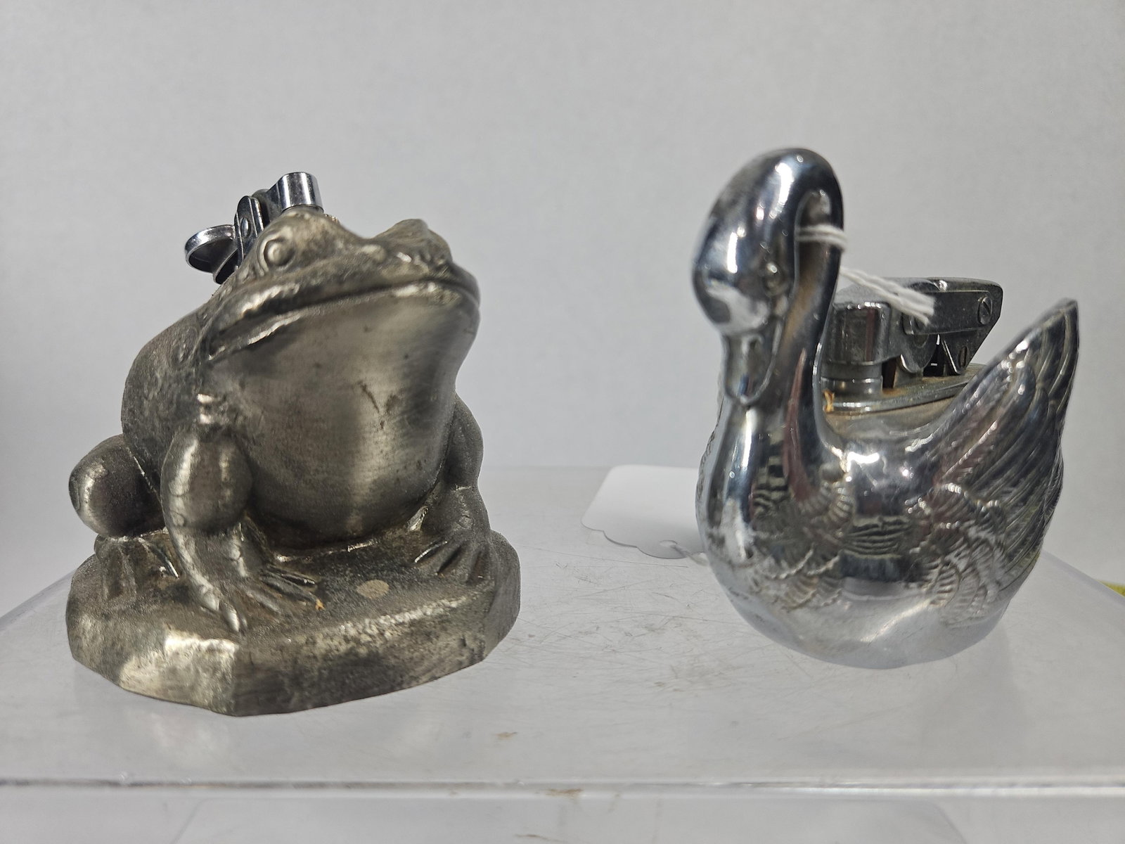 Pair Metal Frog & Swan Table Lighters: This lot comprises two vintage novelty table lighters fashioned in animal forms. The first piece is a cast metal frog situated on an irregular rocky base, finished in a matte grey tone. The back of