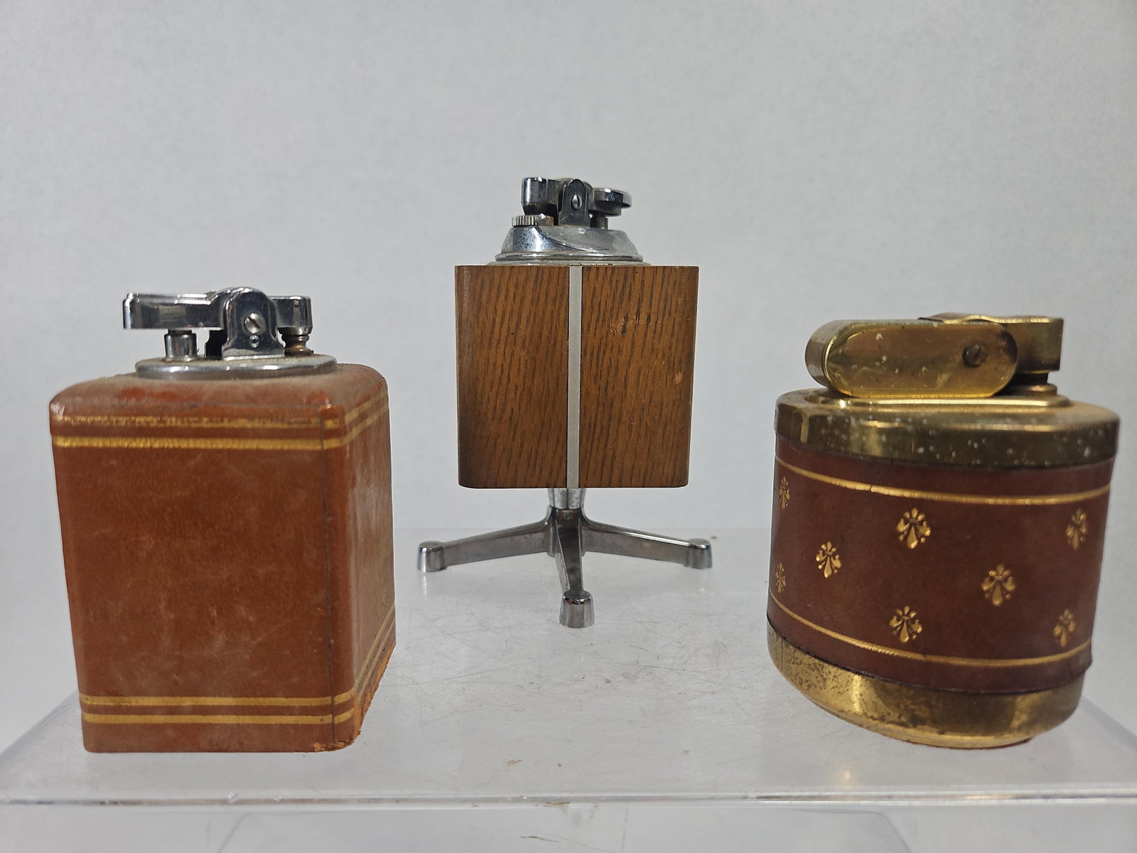 Set 3 Mid-C. Table Lighters w/ Ronson: A trio of vintage mid-century table lighters featuring various materials and decorative styles. The first lighter is a Ronson brand model with a square body wrapped in brown leather, featuring two