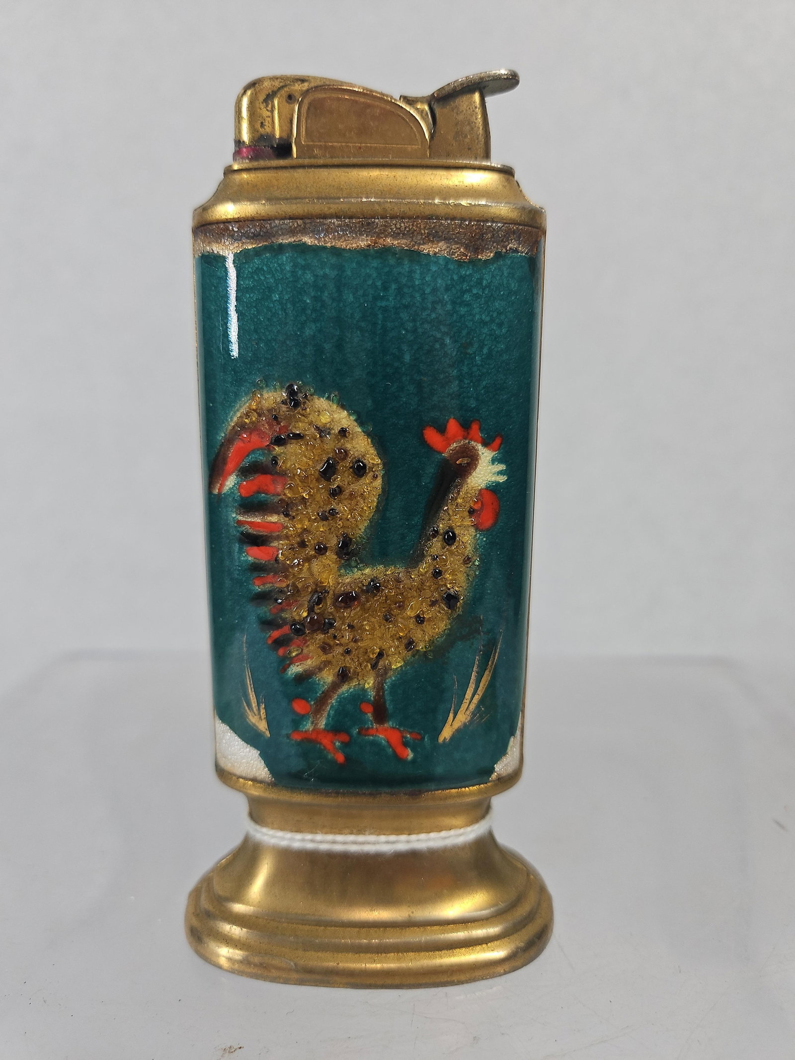 Evans Enamel & Brass Rooster Lighter: This mid-century table lighter features an oval pillar-form body decorated with a dark teal green stippled enamel ground. The front is embellished with a stylized rooster rendered in raised, textured