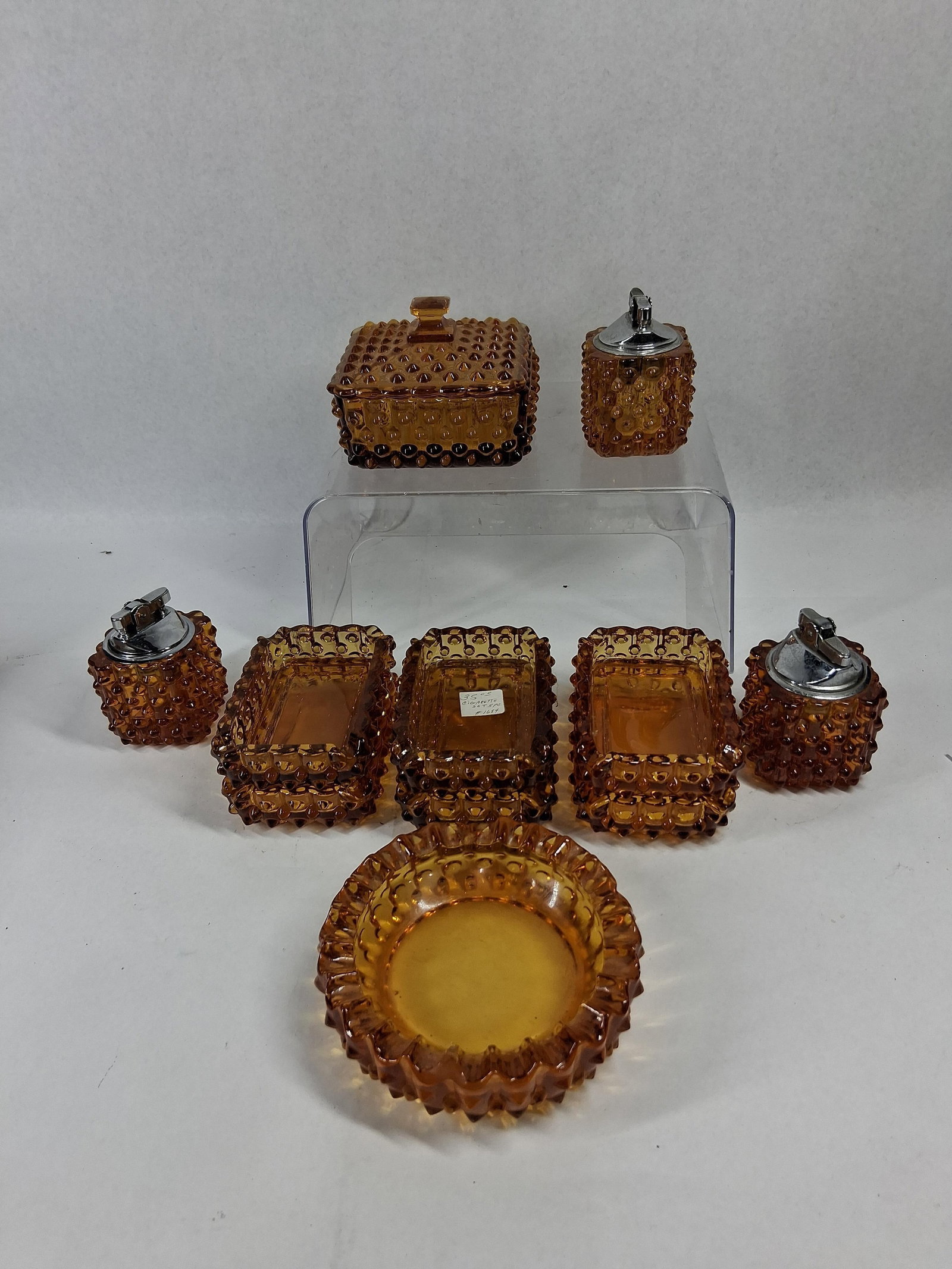 8-Piece Amber Hobnail Glass Smoking Set: This eight-piece smoking set is constructed from transparent amber-colored pressed glass featuring a uniform pointed hobnail pattern throughout. The set includes a square lidded cigarette box, three