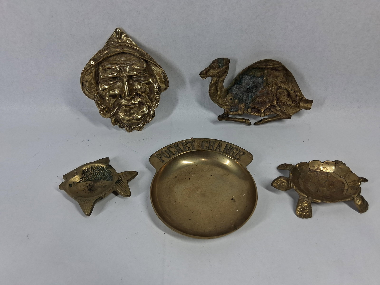 5 Vtg Cast Brass Novelty Trays & Dishes: An assortment of five vintage cast brass novelty items designed as trinket dishes, ashtrays, or decorative plaques. The collection includes a circular tray with a raised rim featuring the text POCKET