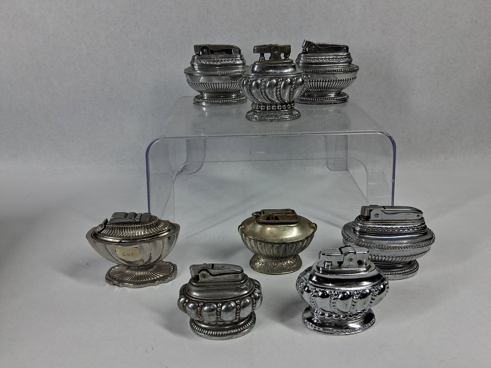 ASR & Omsco Silver-Plate Table Lighters: A collection of vintage silver-plated table lighters featuring various decorative motifs and mechanical styles. The group includes footed lighters with designs such as gadrooning, beadwork, fluting, a