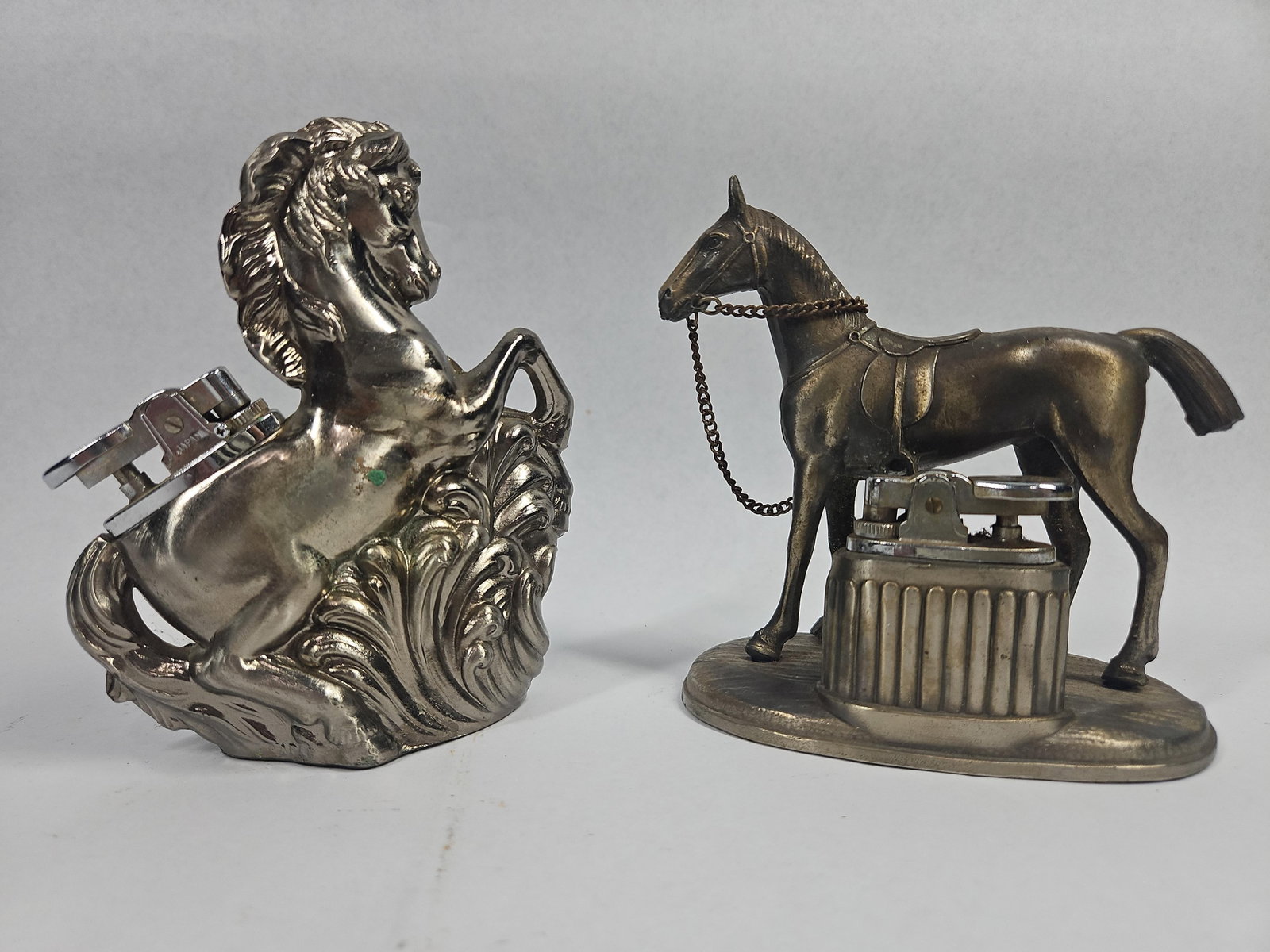 Pair Cast Metal Horse Table Lighters: This lot consists of two cast metal equestrian-themed table lighters. The first piece features a standing saddled horse with a metal chain bridle mounted on an oval base next to a fluted cylindrical