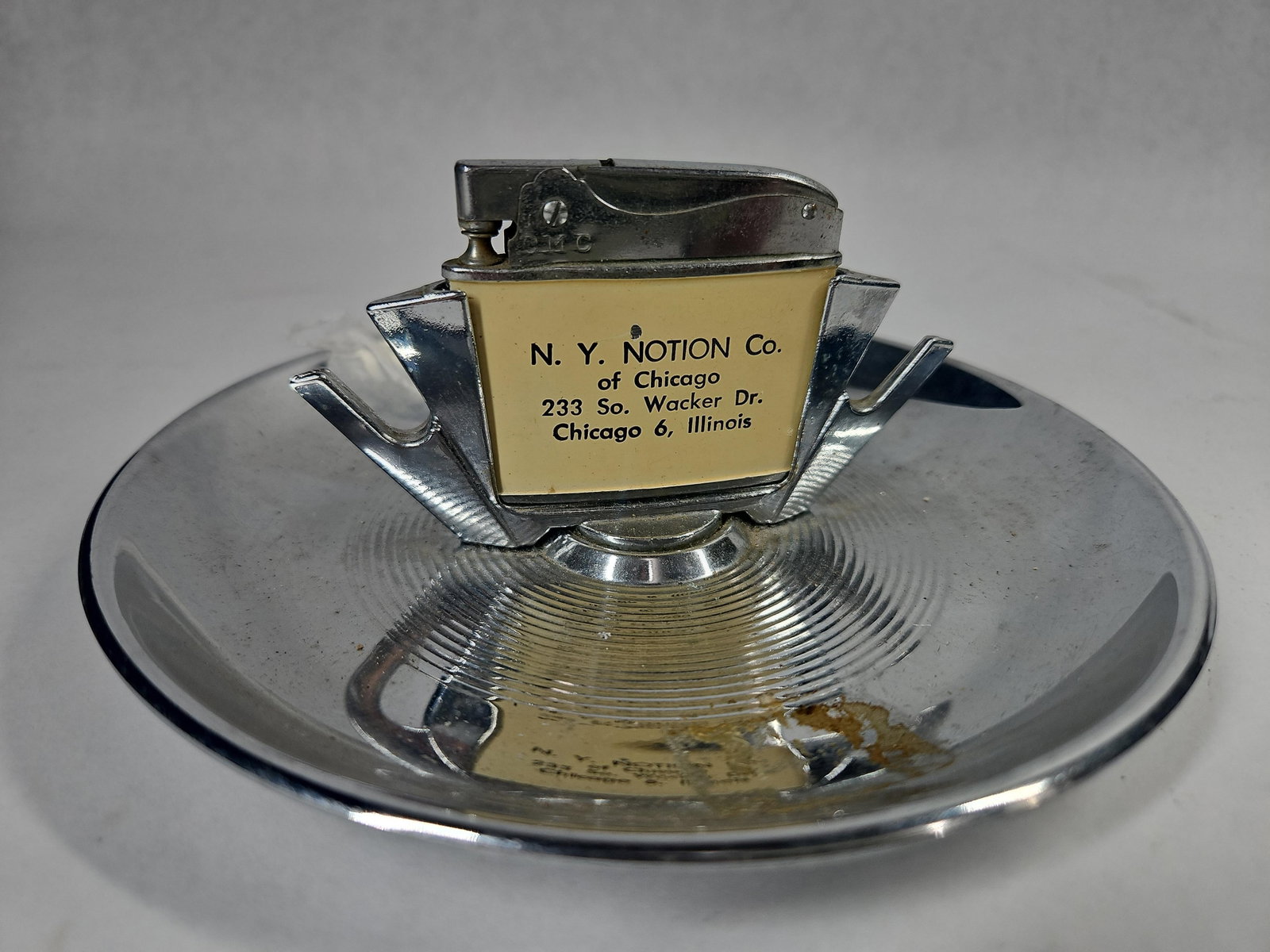 N. Y. Notion Co. Chrome Lighter/Ashtray: A vintage chrome-plated metal desk lighter with an integrated circular ashtray. The central lighter housing features an inset cream-colored plastic panel printed with black text reading N. Y. NOTION