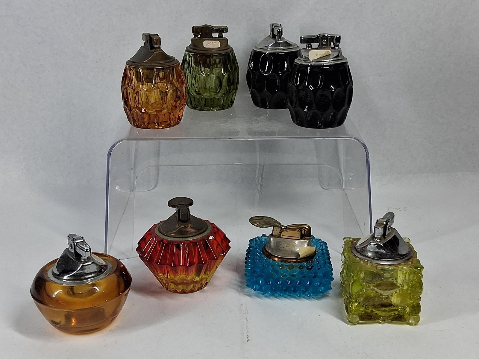 8 Assorted Vtg Glass Table Lighters: An assortment of eight glass-bodied table lighters featuring various pressed and faceted designs. The collection includes four thumbprint-patterned barrel-form lighters in amber, emerald green, and