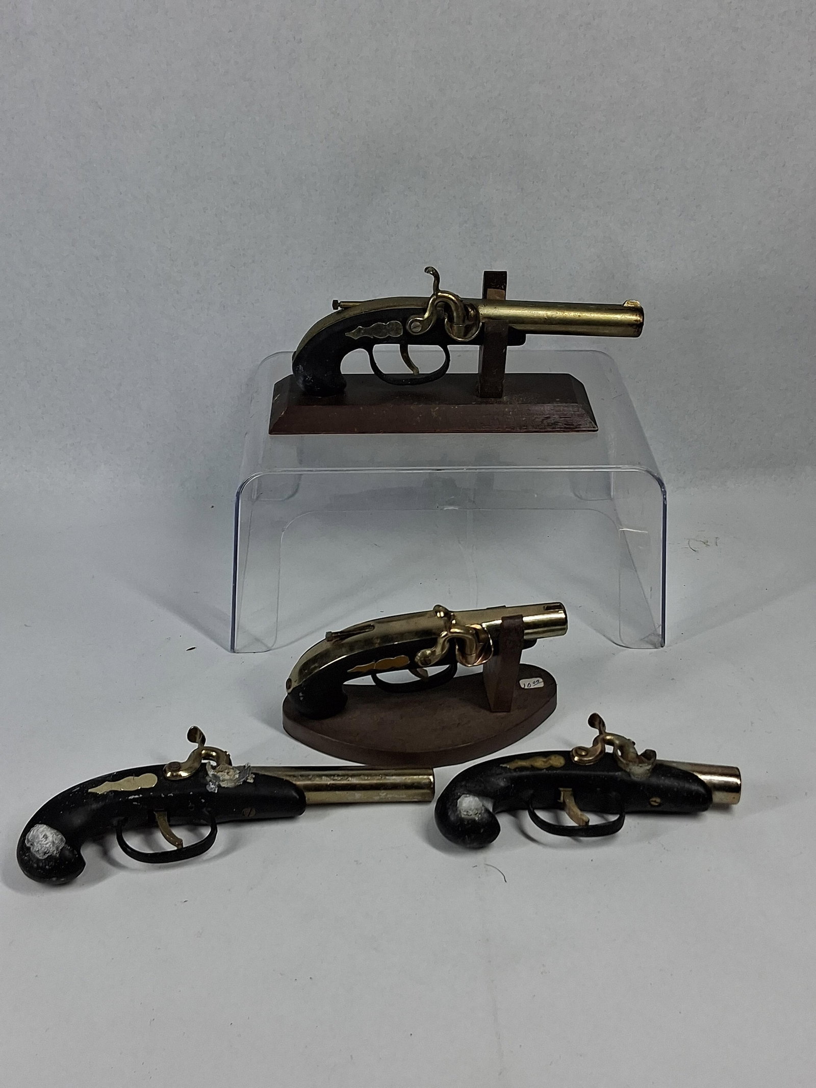 Percussion-Style Pistol Table Lighters: An assortment of miniature decorative pistols and dark-stained wooden display stands. Each piece is modeled after a percussion-style firearm, featuring brass-toned metal barrels and hammer assemblies