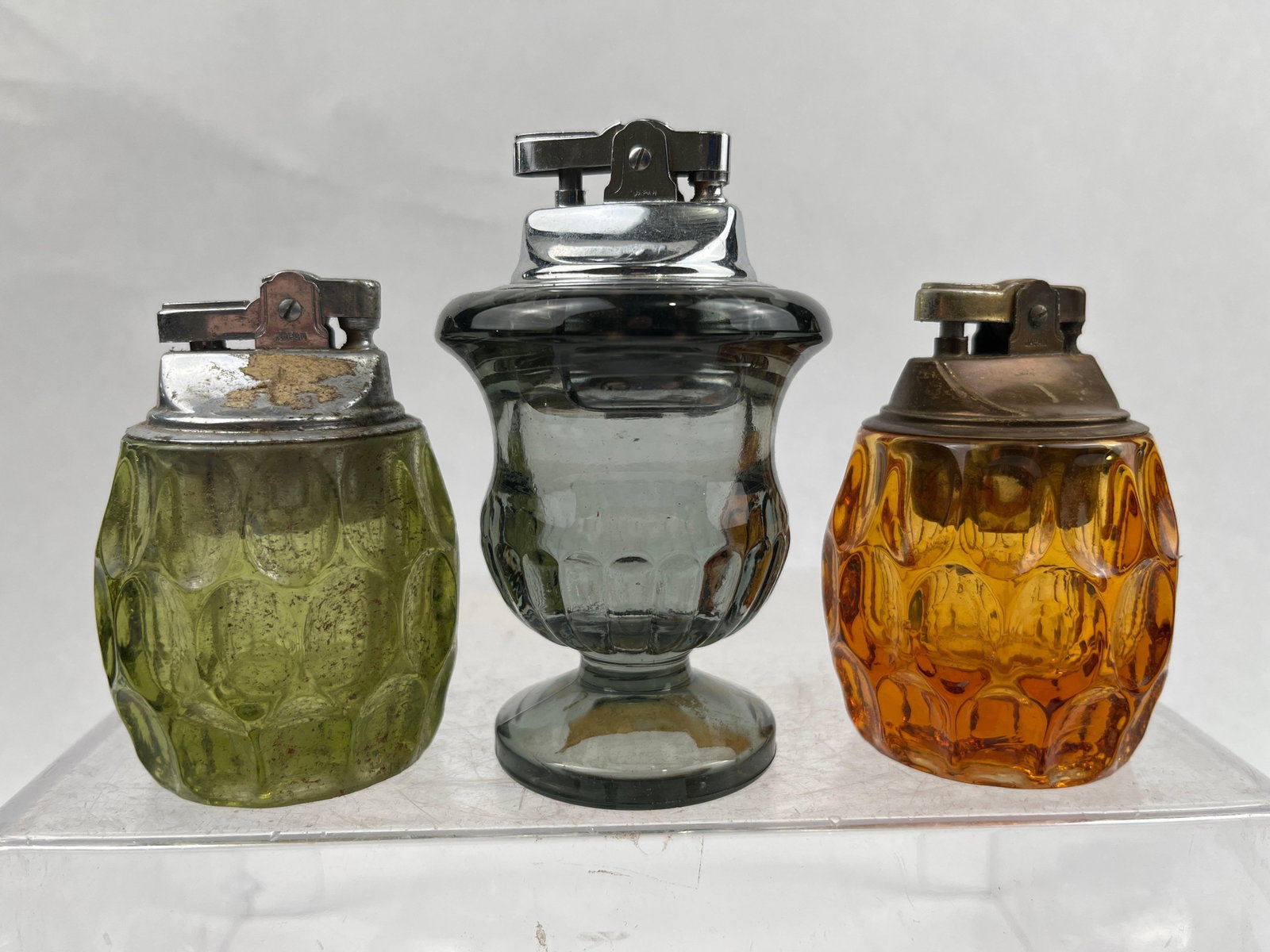 King Japan Pressed Glass Table Lighters: An assortment of mid-century modern table lighters featuring pressed glass bases in various colors including olive green, amber, smoke grey, and dark cobalt blue. The collection includes vessels with 