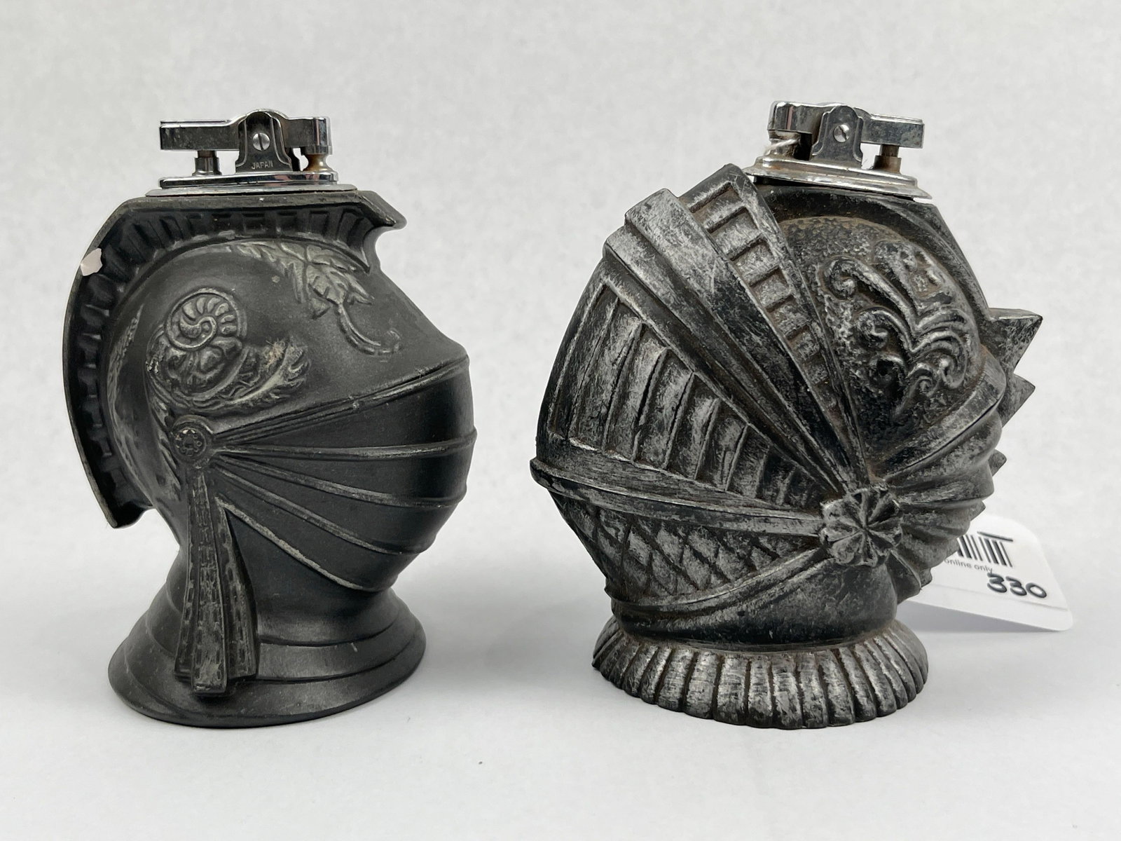 Pair Japanese Knight Helmet Lighters: A pair of vintage cast metal table lighters modeled as medieval knight helmets. The first helmet features a closed-face design finished in a matte charcoal patina with embossed scroll and foliate moti