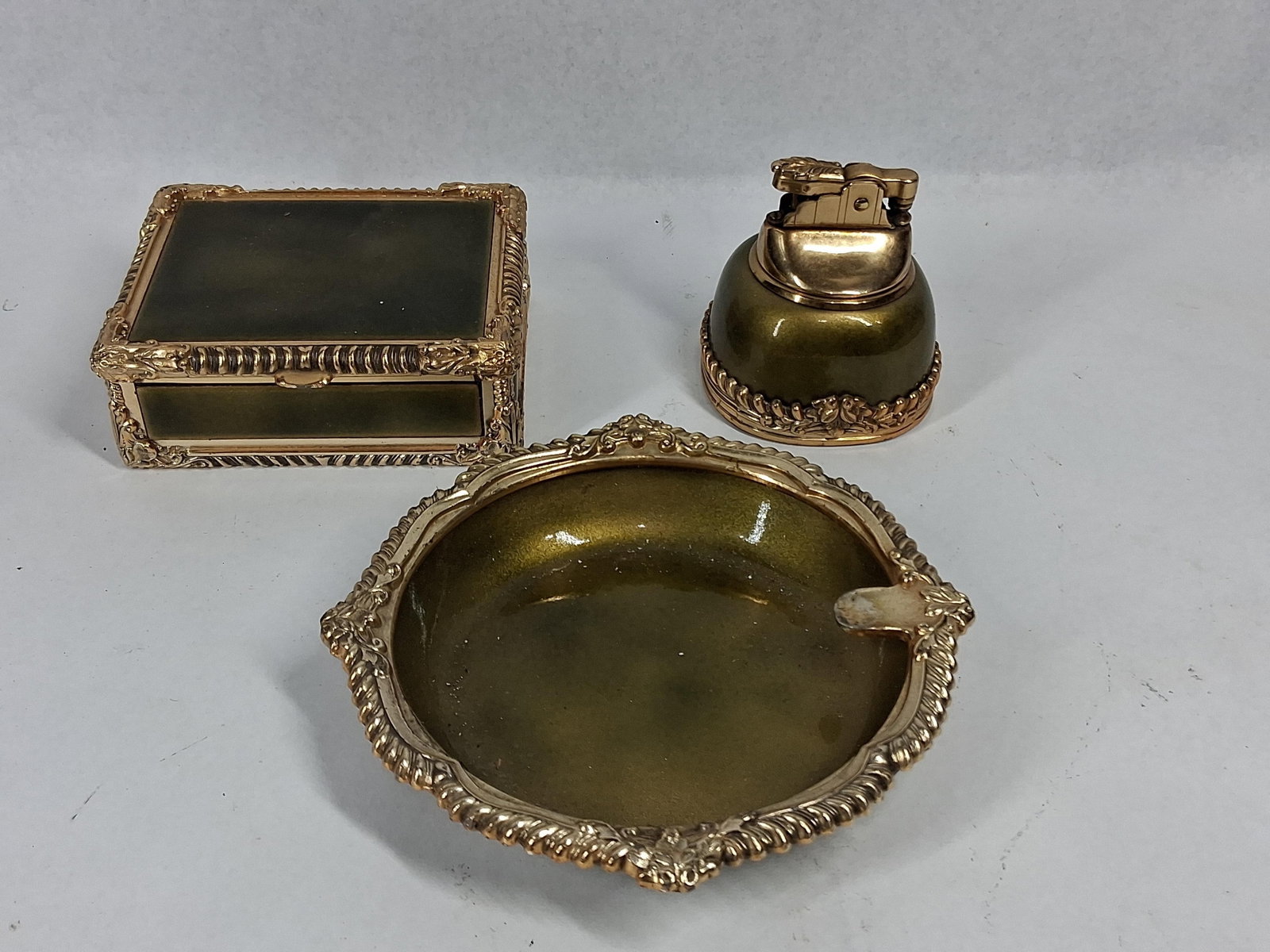Royal Bovano Enameled Metal Smoking Set: A three-piece smoking set comprising a rectangular hinged box, a table lighter, and a circular ashtray. The pieces are constructed from gold-tone cast metal featuring ornate borders with shell and scr