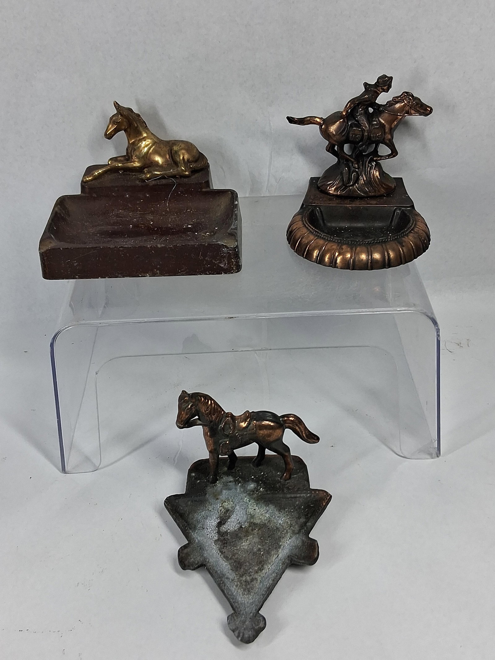 Trio Phil. & Rodeo Metal Horse Ashtrays: An assortment of three vintage cast metal ashtrays with equine decorative elements. The first is a rectangular two-tiered ashtray supporting a figure of a recumbent foal. The horse figure features a