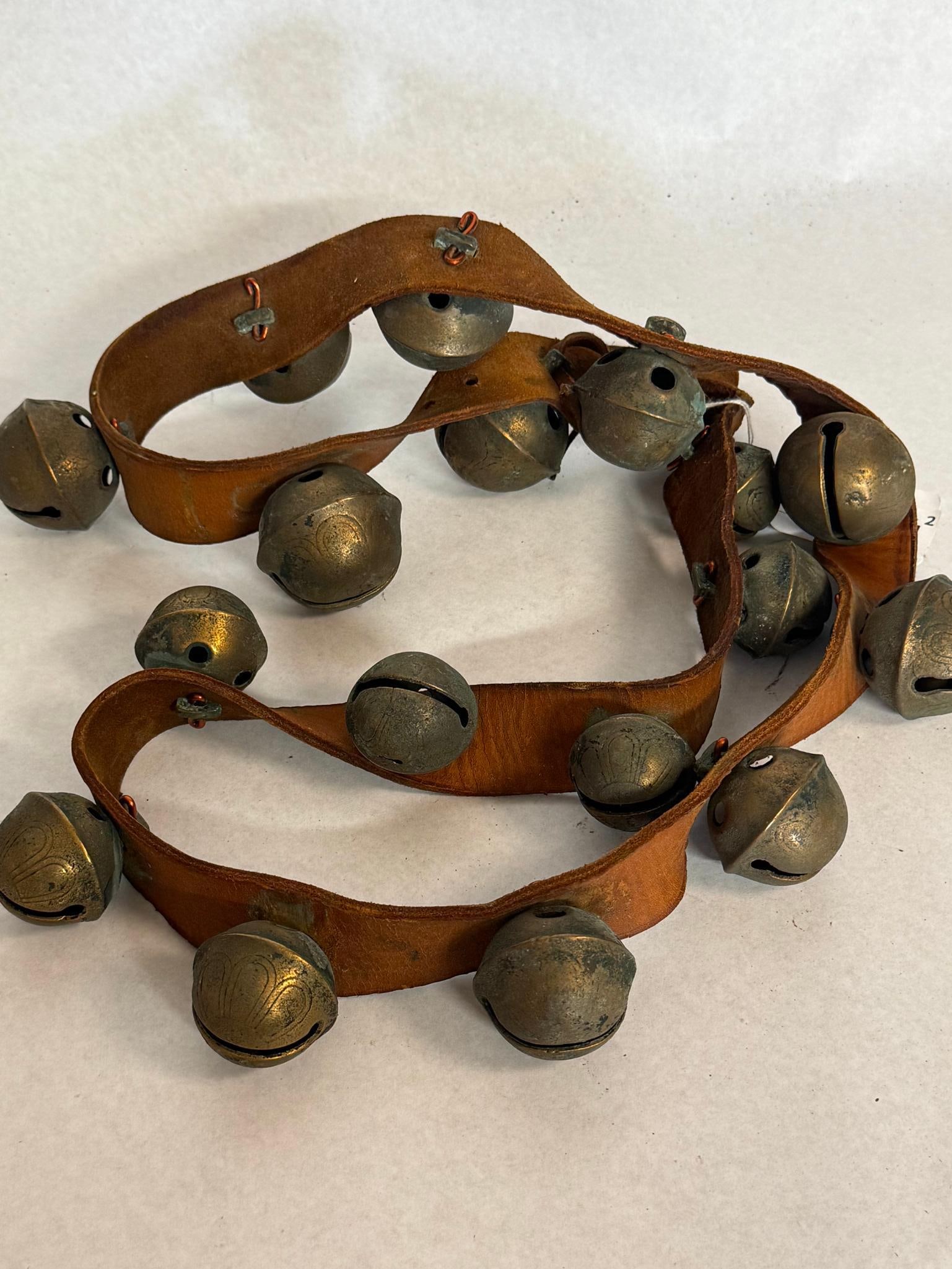 Antique Cast Brass Petal Sleigh Bells: A set of cast brass or bronze crotal bells, commonly referred to as sleigh bells, mounted on a single length of brown leather. The bells are constructed with a horizontal slit at the base terminating
