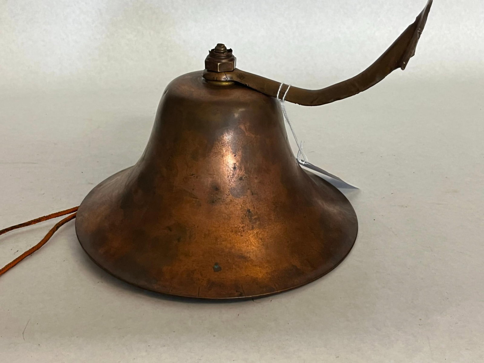 Wrought Copper Ship Bell w/ Brass Arm: This wall-mounted bell is constructed from sheet copper featuring a wide flared rim and a visible vertical seam, indicating a hand-wrought fabrication process. The bell is equipped with a curved, cast