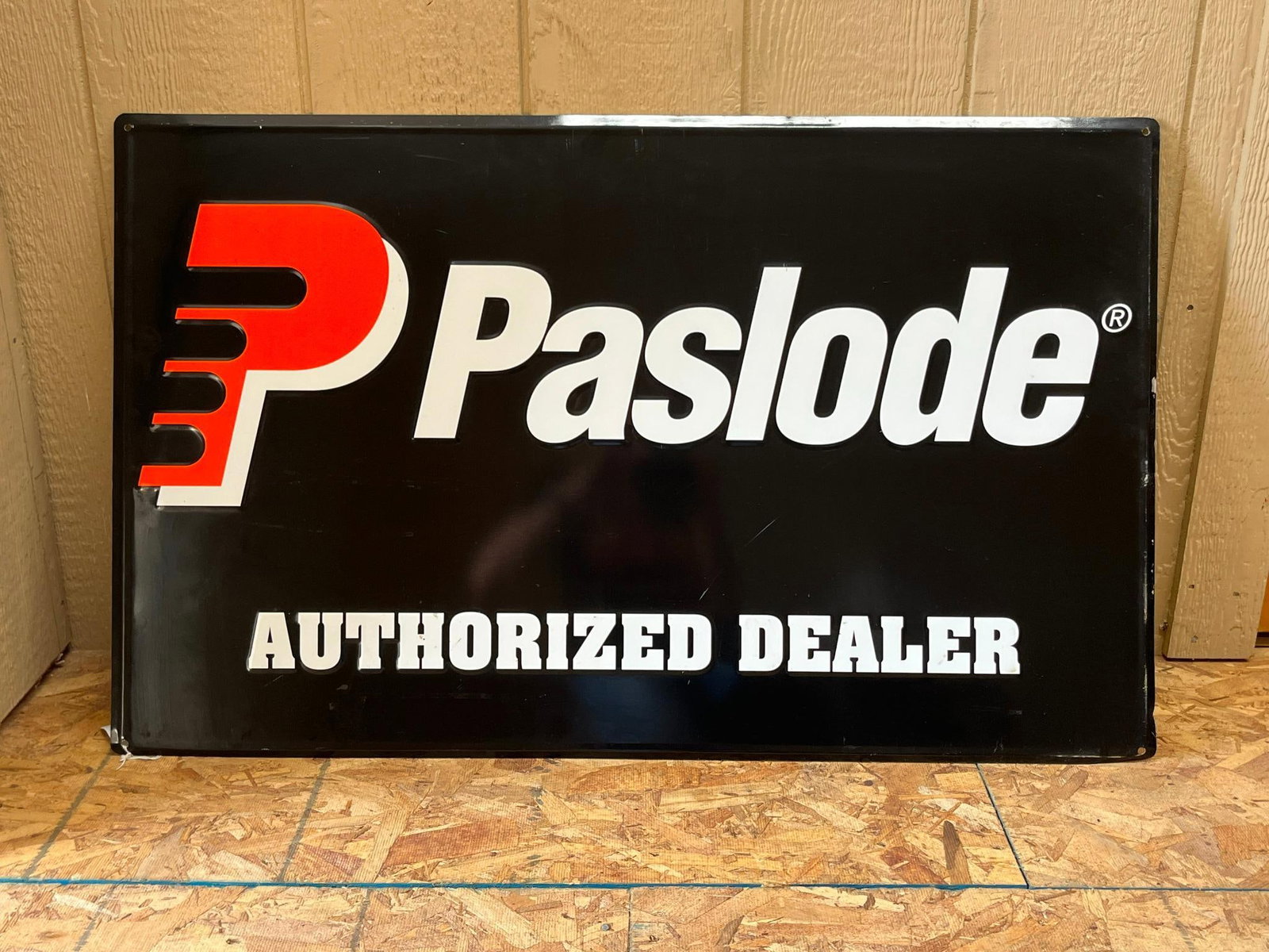 Paslode Embossed Metal Advertising Sign: A single-sided, rectangular embossed metal advertising sign for the tool brand Paslode. The Paslode logo on the left is composed of a stylized red and white letter P with horizontal bars. To the right