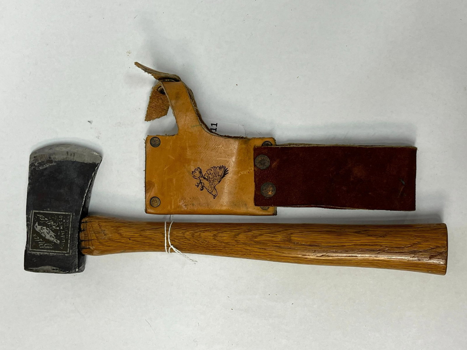 Kelly Axe Black Raven Hatchet w/ Sheath: An American-made forged steel hatchet featuring a wood handle and a coordinating two-tone leather sheath. The face of the axe head displays a rectangular etched mark with a graphic of a bird and