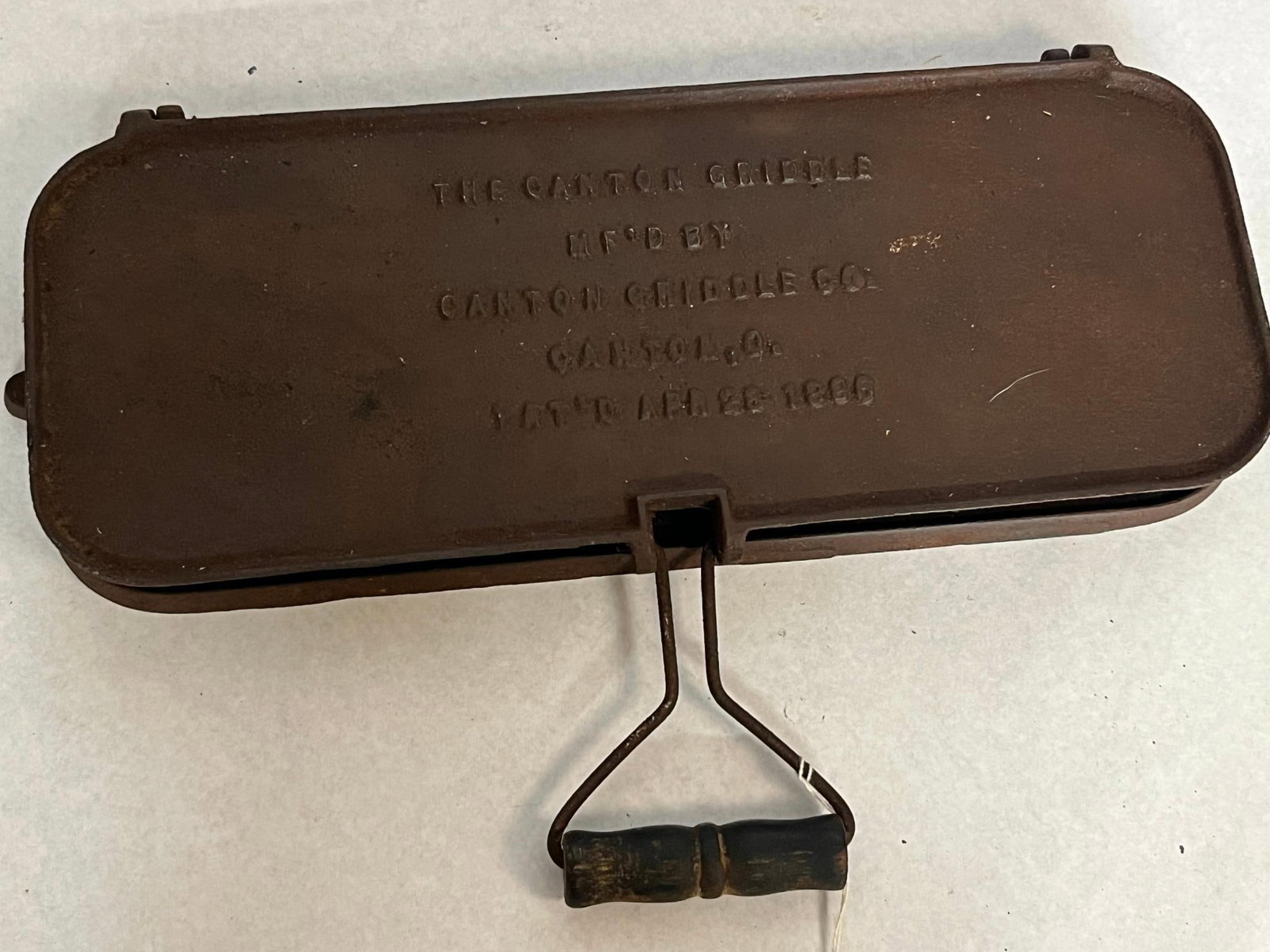Canton Griddle Co Iron 3-Cake Griddle: An American cast iron flip griddle featuring two rectangular plates joined by hinges. It is equipped with a steel wire handle featuring a turned wooden grip. An embossed mark in raised lettering on