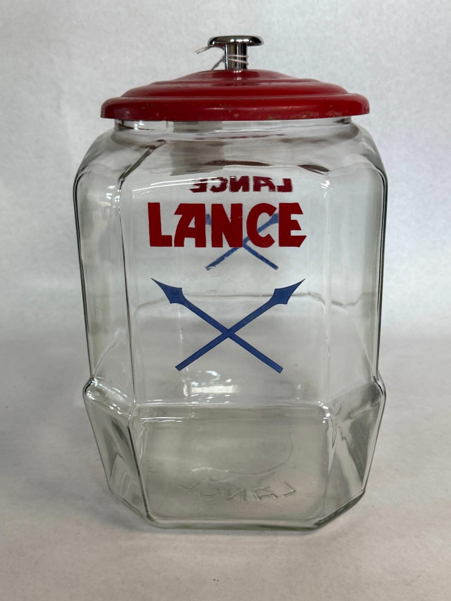 Lance 8-Sided Glass Cracker Jar w/ Lid: A clear glass countertop snack display jar produced for the Lance Packing Company. Two opposing sides of the jar are decorated with the company logo, featuring the word LANCE in red block lettering po