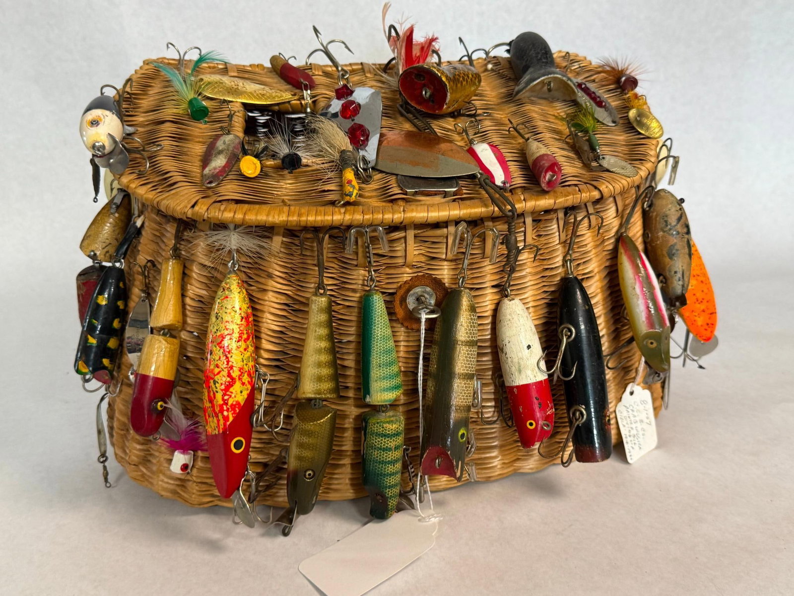 Gibbs & Yakima Lures & Wicker Creel: An assortment of antique and fishing tackle displayed on a split-willow wicker creel. The wicker basket is constructed from hand-woven split willow with a hinged lid and a metal pin closure at the