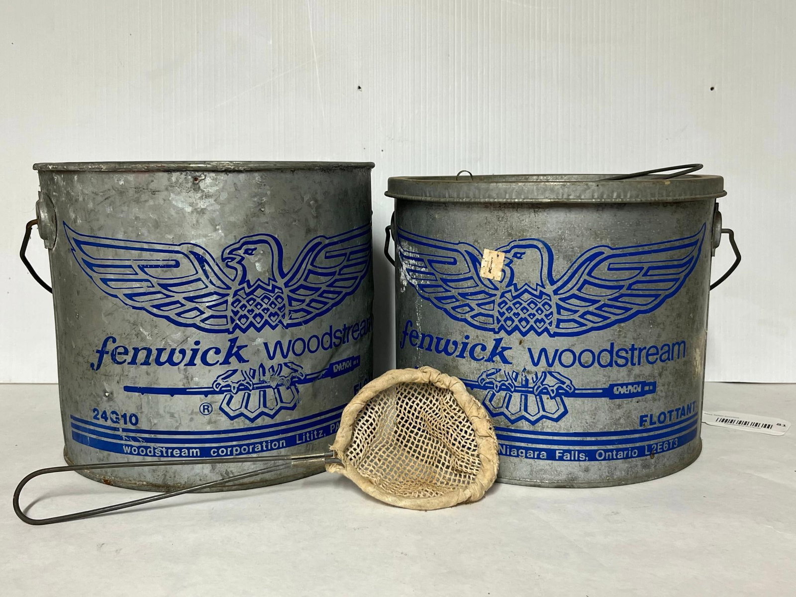 Fenwick Woodstream Minnow Buckets & Net: A set of fishing equipment featuring two galvanized metal bait buckets and a small hand net. Both buckets are decorated with blue screen-printed graphics depicting an eagle logo above the text