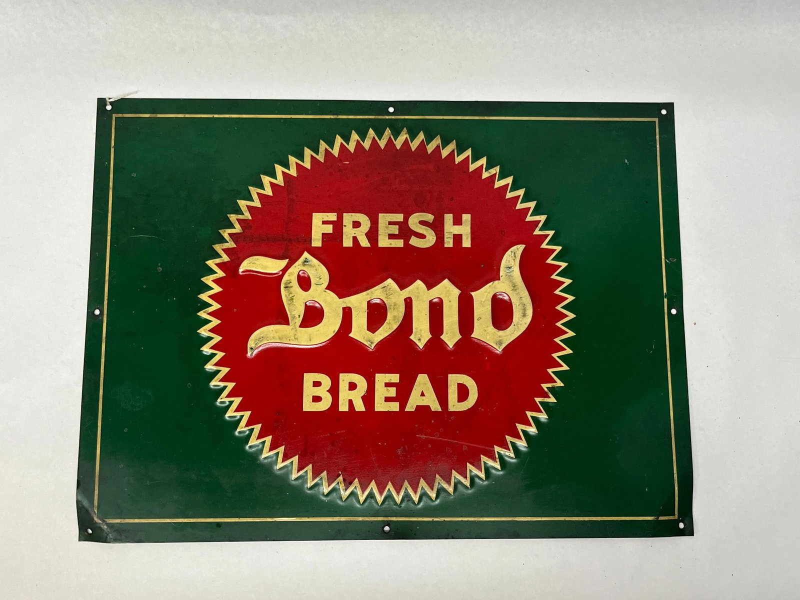 Bond Bread Embossed Tin Sign, 20th C: This rectangular embossed tin advertising sign features a dark green background with a central red saw-toothed sunburst graphic. The sign is constructed from sheet metal and includes eight