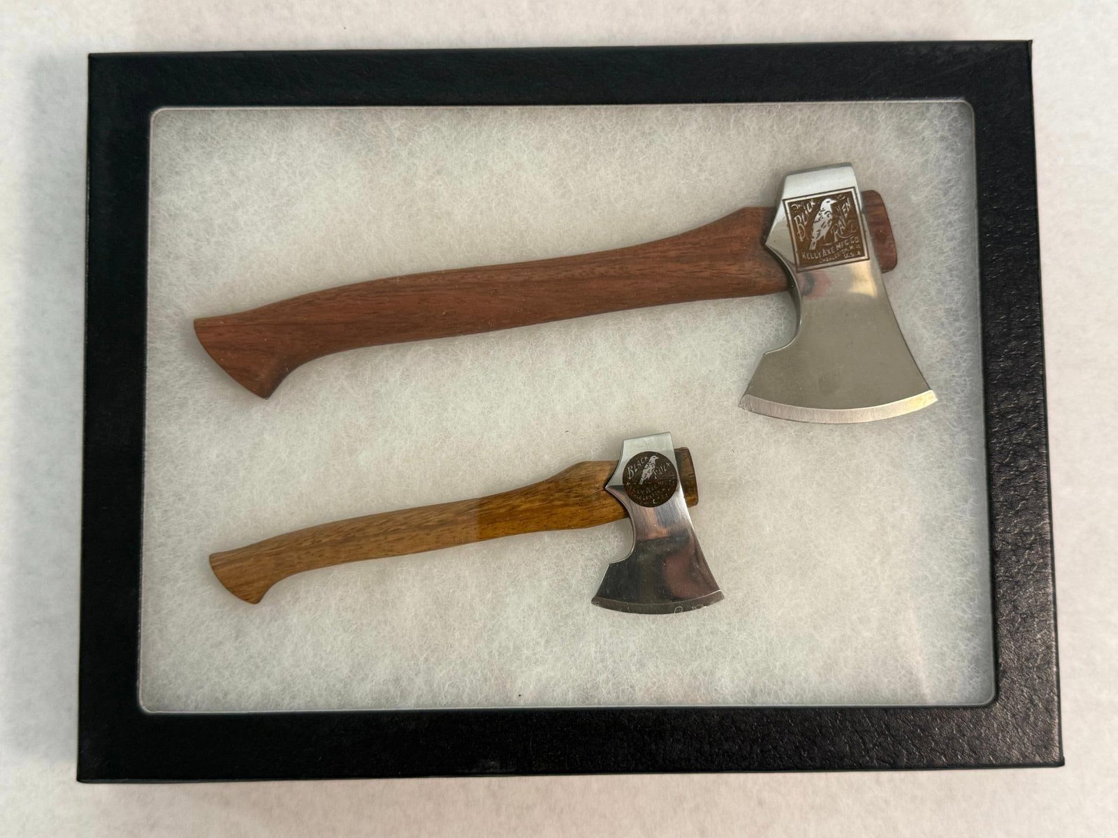 Pair Kelly Axe Black Raven Tribute Axes: A pair of miniature axes featuring highly polished steel heads and carved hardwood handles. The larger axe features a dark reddish-brown wood handle, while the smaller axe is fitted with a lighter gol