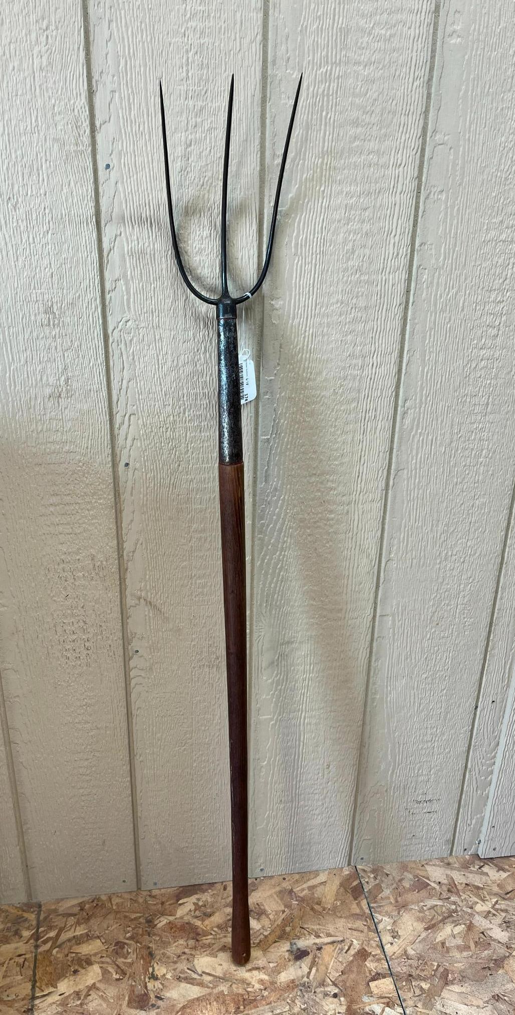 Three-Tine Forged Steel & Wood Hay Fork: A three-tine pitchfork, also known as a hay fork, designed for agricultural use in moving loose hay, straw, or garden debris. The tool features a forged steel head with three long, upwardly curved