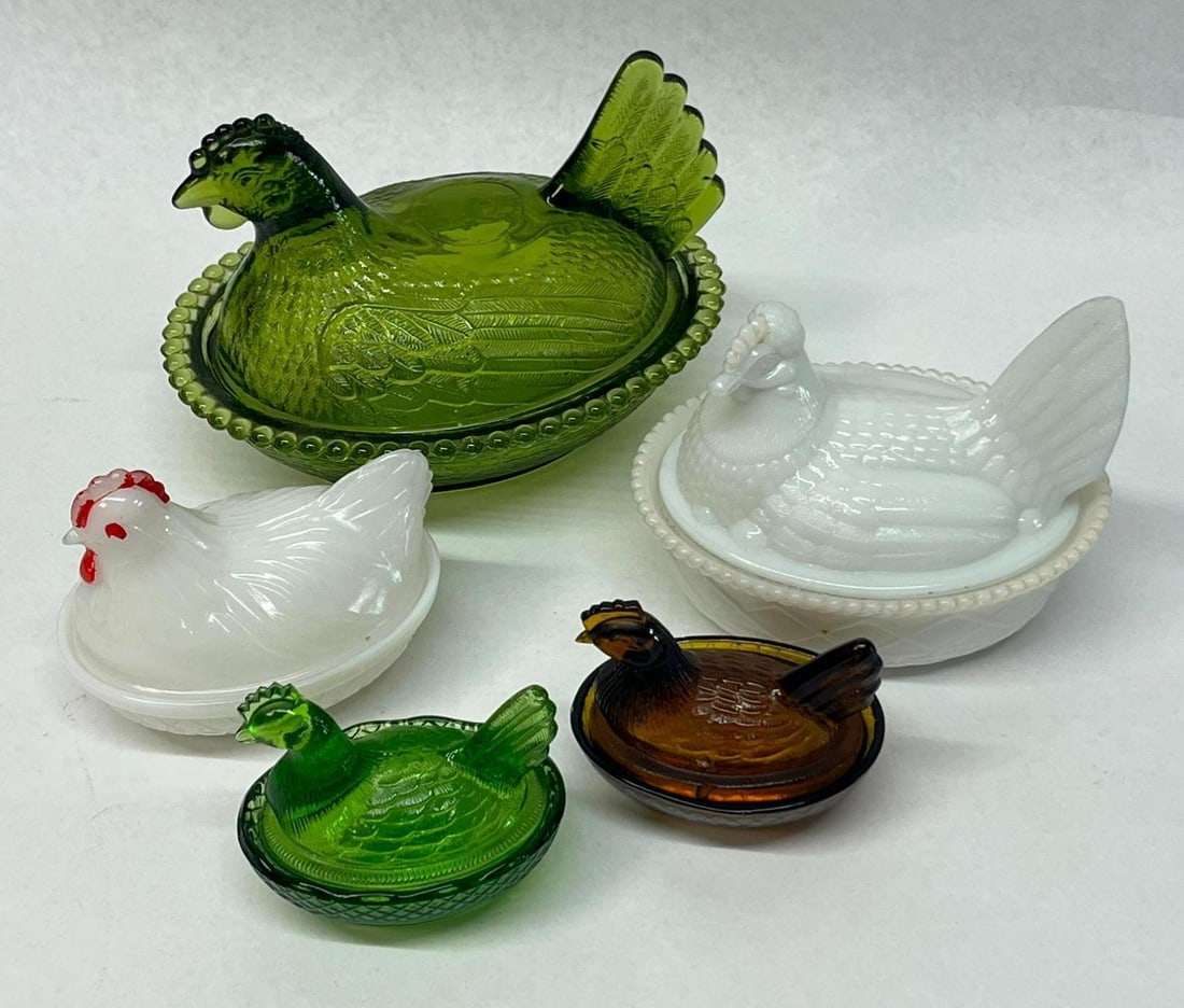 Set 5 Glass Hen on Nest Covered Dishes: This collection consists of five glass hen on nest covered dishes in various sizes and colors. The largest piece is a transparent deep green pressed glass dish featuring a hen lid with detailed