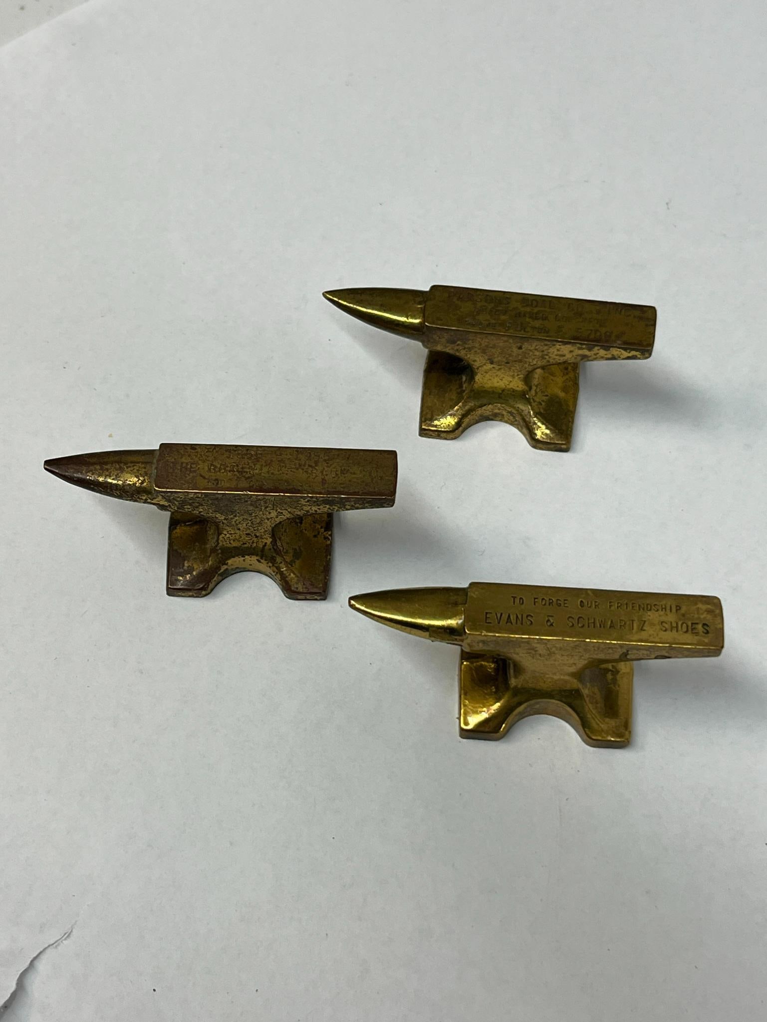 3 Cast Brass Advertising Anvils: A trio of mid-20th-century promotional miniature anvils cast from metal with a brass-toned finish. The first anvil bears an impressed inscription on the top face that reads TO FORGE OUR FRIENDSHIP