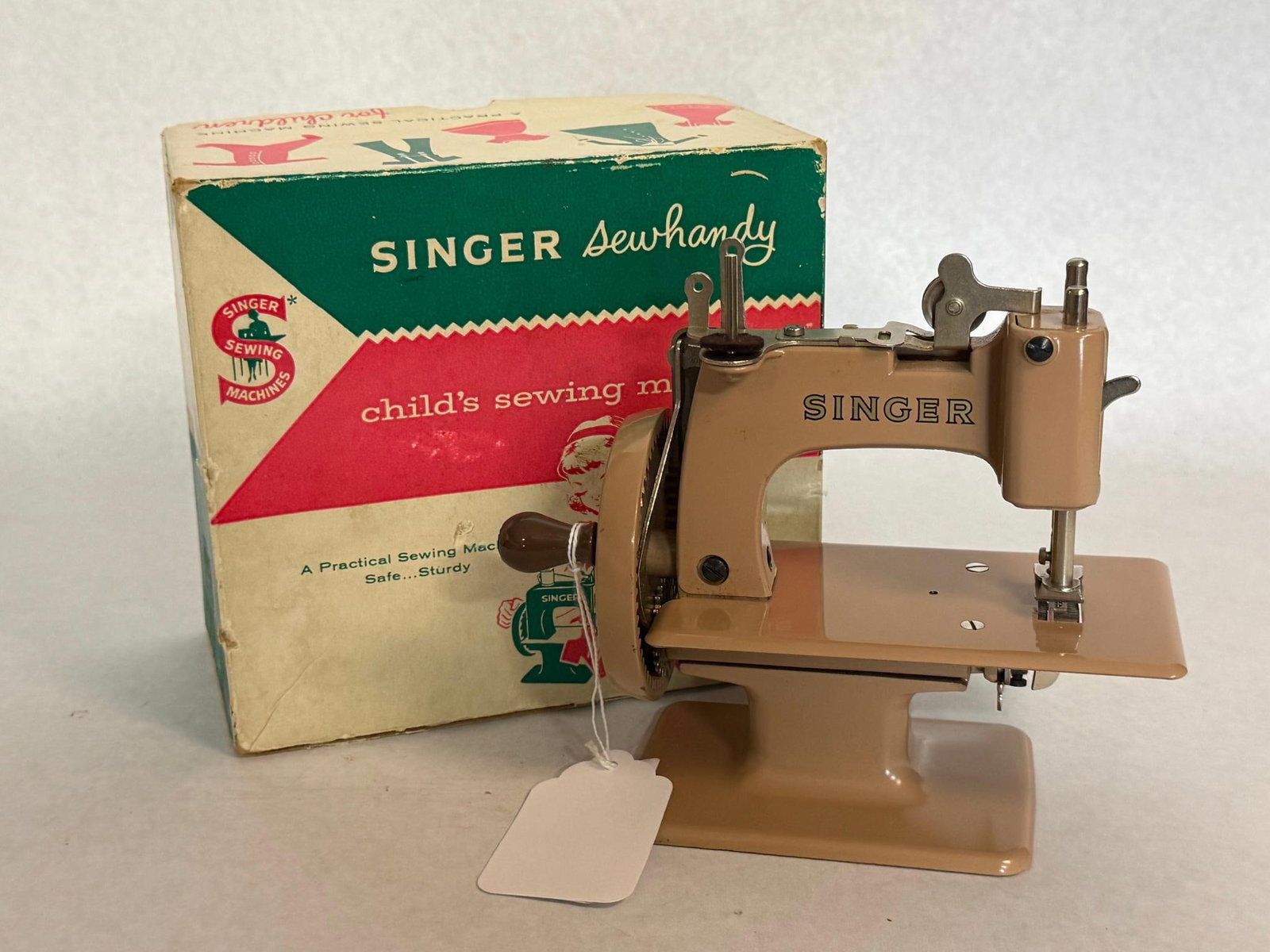 Singer Sewhandy Model 20 Sewing Machine: A 1950s-era American child hand-crank sewing machine featuring a die-cast metal body and base with a high-gloss beige painted finish. A circular metal badge in black and gold tones is affixed to the