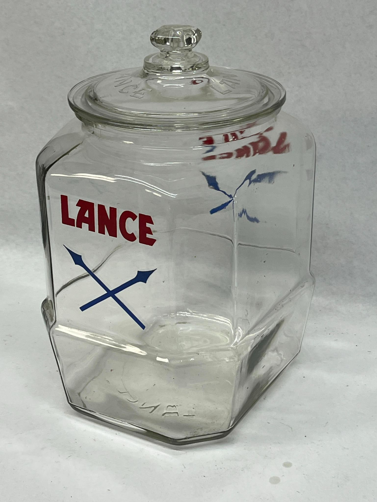 Lance Eight-Sided Glass Cracker Jar: A mid-20th-century American clear glass counter display jar for the Lance snack company. The exterior is decorated on two sides with the company brand name LANCE in red block letters above a blue