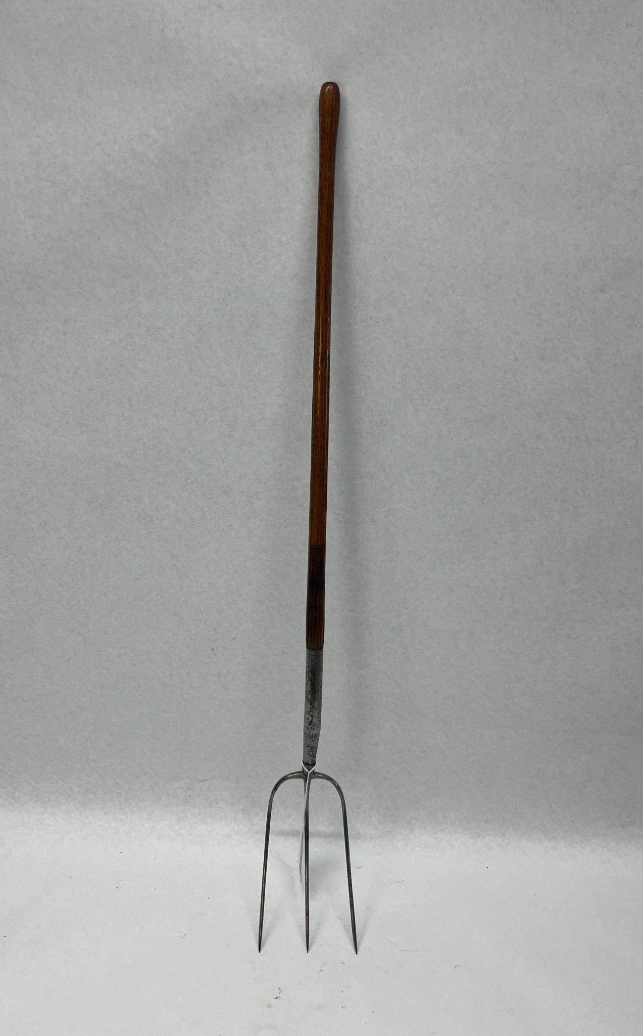 Three-Tine Agricultural Wood Pitchfork: A agricultural hand tool comprising a forged metal head with three curved tines and a long wooden handle. The metal head is constructed with three slender tines attached to a central bridge that