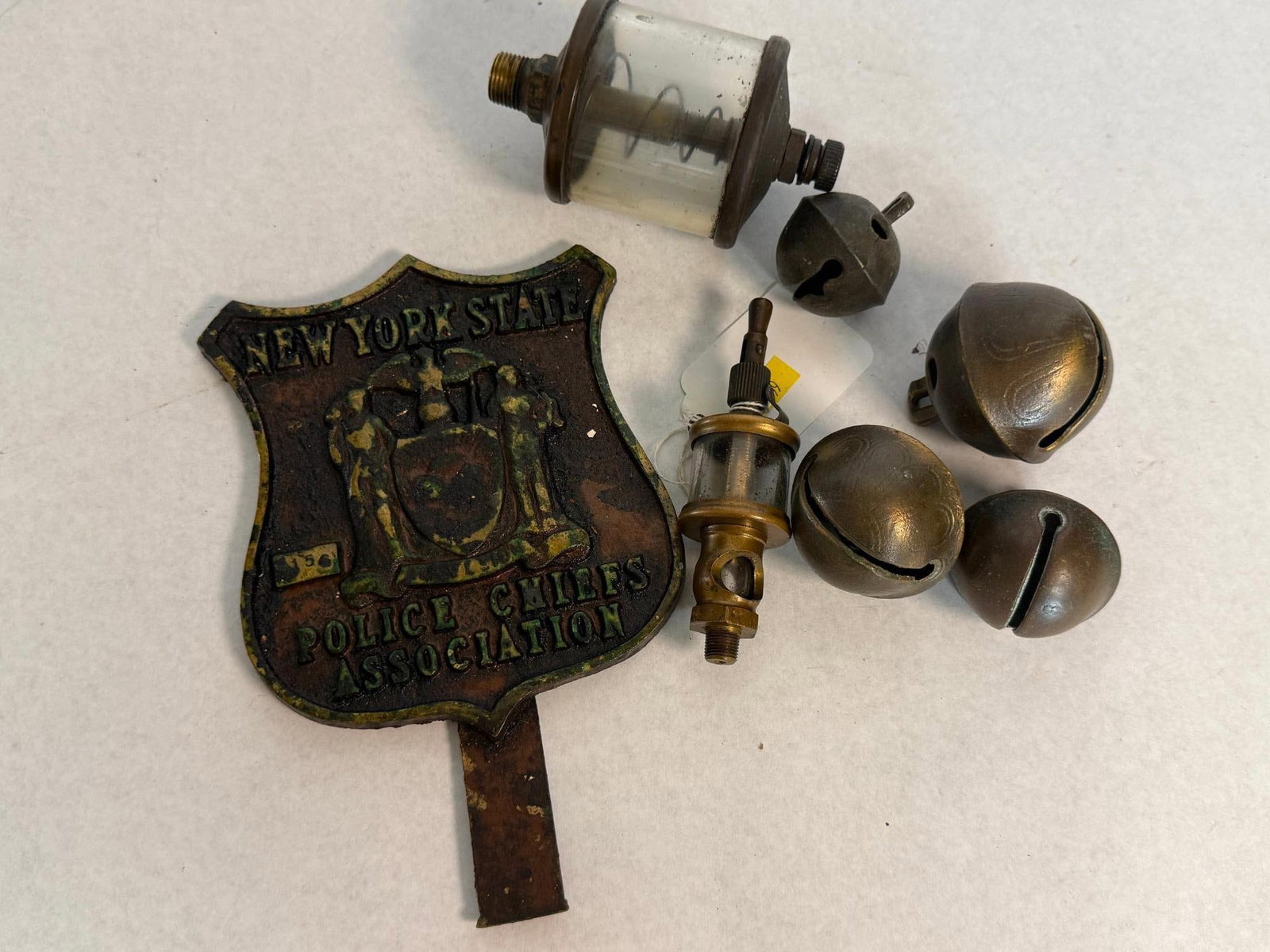 Auto & Ind. Collectibles w/ NYS Badge: A collection of automotive and industrial artifacts including one radiator badge, two drip feed oilers, and four crotal bells. The face features the New York state seal in relief between the text NEW