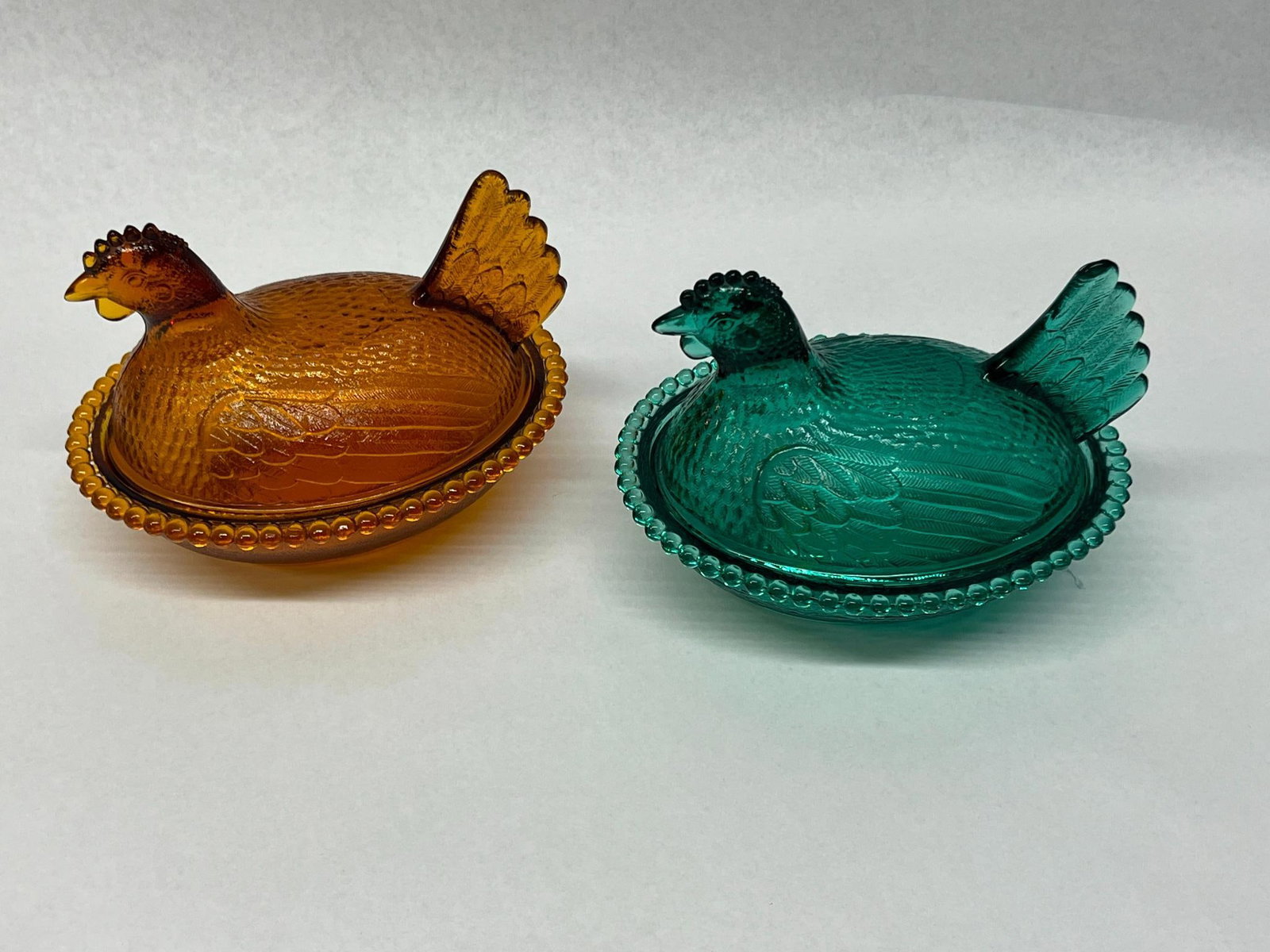 Pair Amber & Teal Hen-on-Nest Dishes: A pair of two-part pressed glass covered dishes in the form of nesting hens. This lot includes one dish in a deep amber color and one in a vibrant teal glass. The bases feature a textured basketweave 