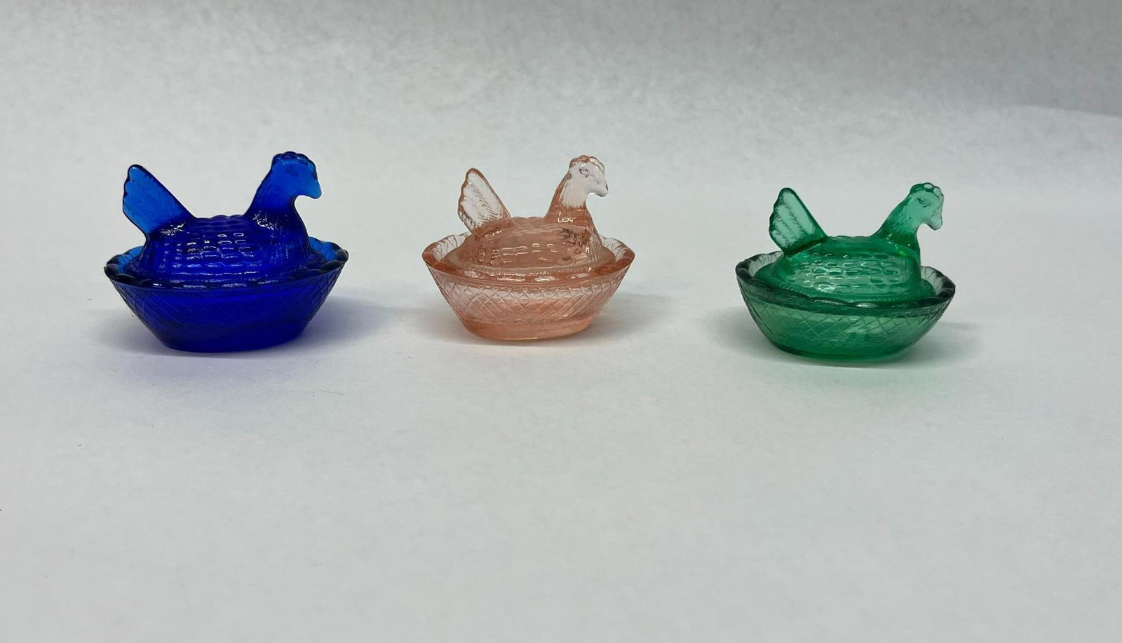 Trio Pressed Glass Hen on Nest Dishes: A trio of miniature covered dishes in the form of a hen on a nest, crafted from pressed transparent glass. The collection includes three distinct colors: cobalt blue, transparent pink, and transparent