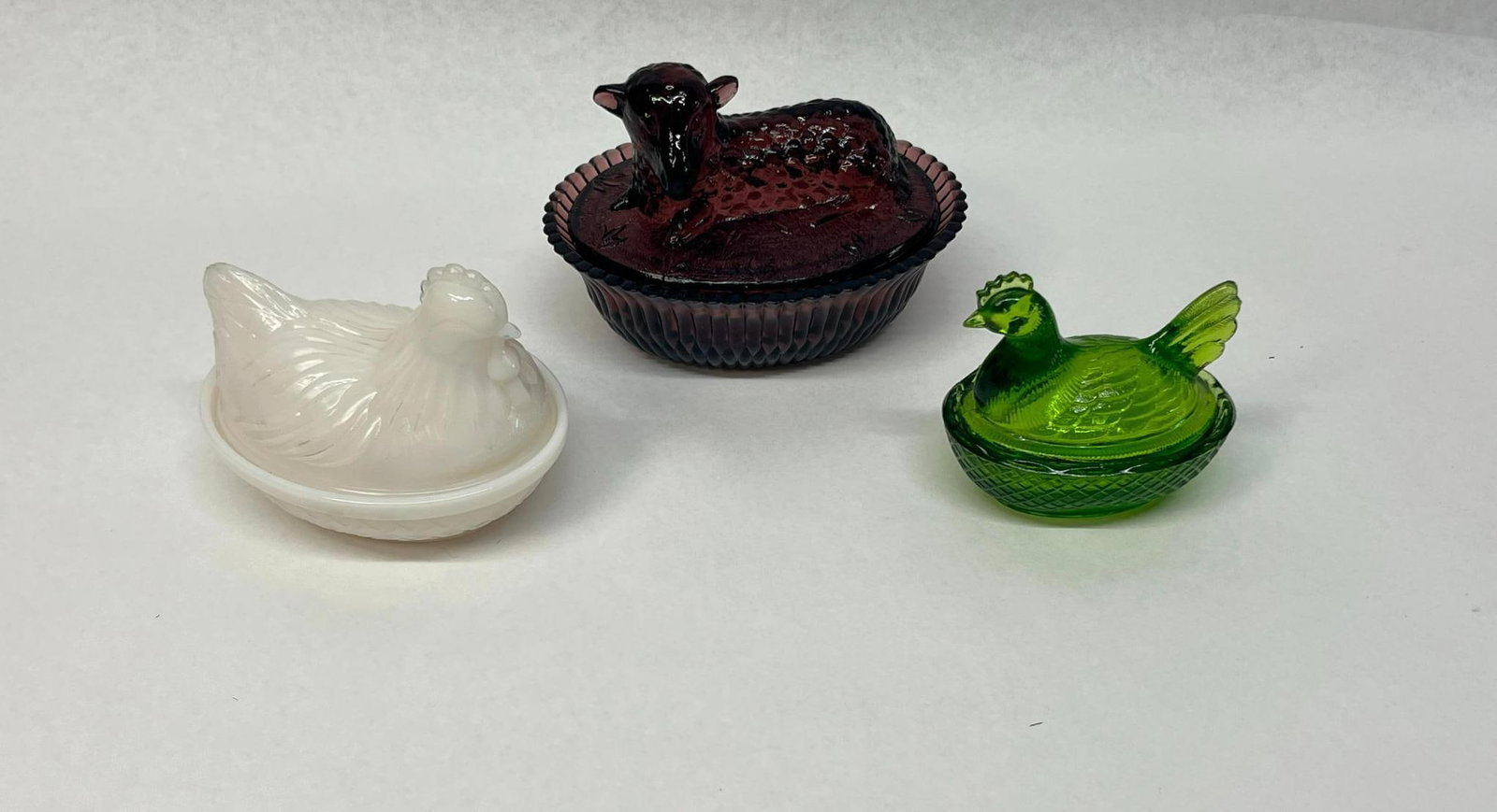 Trio Pressed Glass Animal Dishes: This lot features three pressed glass covered animal dishes, commonly used for trinkets or candy. The first is a deep amethyst glass dish with a lid depicting a recumbent lamb on a ground of molded