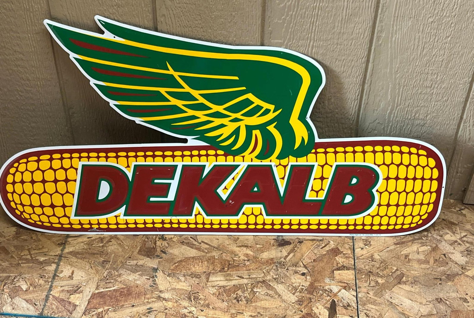 Press Sign Co DeKalb Seed Corn Sign 73: A embossed metal advertising sign for DeKalb, featuring the company logo known as the winged ear of corn. The sign is constructed from a single sheet of metal with a painted finish. A line of small