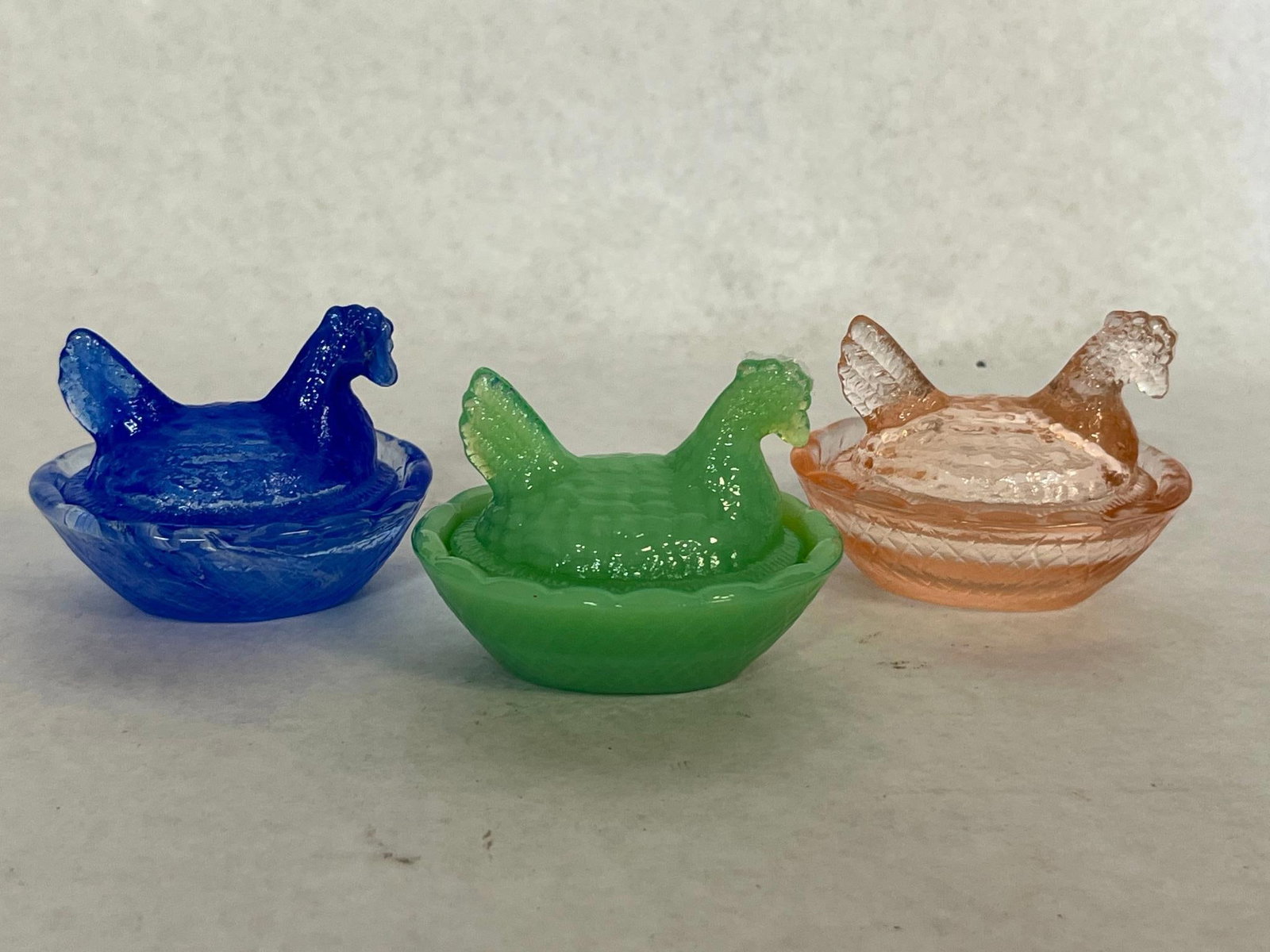 Trio Glass Hen on Nest Covered Dishes: A group of three miniature molded glass covered dishes, each designed as a nesting hen sitting on a basket-form base. The collection includes one dish in translucent cobalt blue slag glass with white