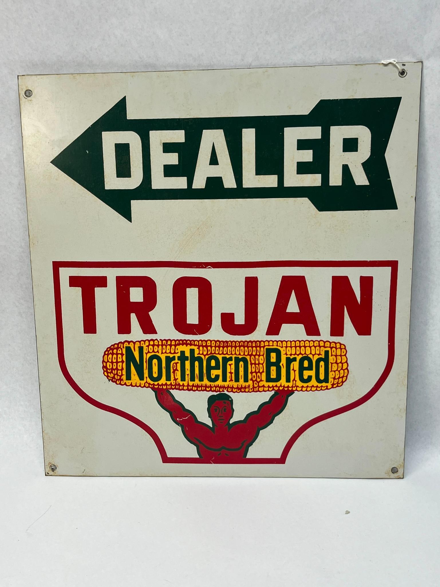Trojan Northern Bred Dealer Metal Sign: A painted metal advertising sign for Trojan Northern Bred seed corn. Below the text is a stylized red muscular figure holding a large yellow ear of corn aloft. No maker marks or manufacturer labels