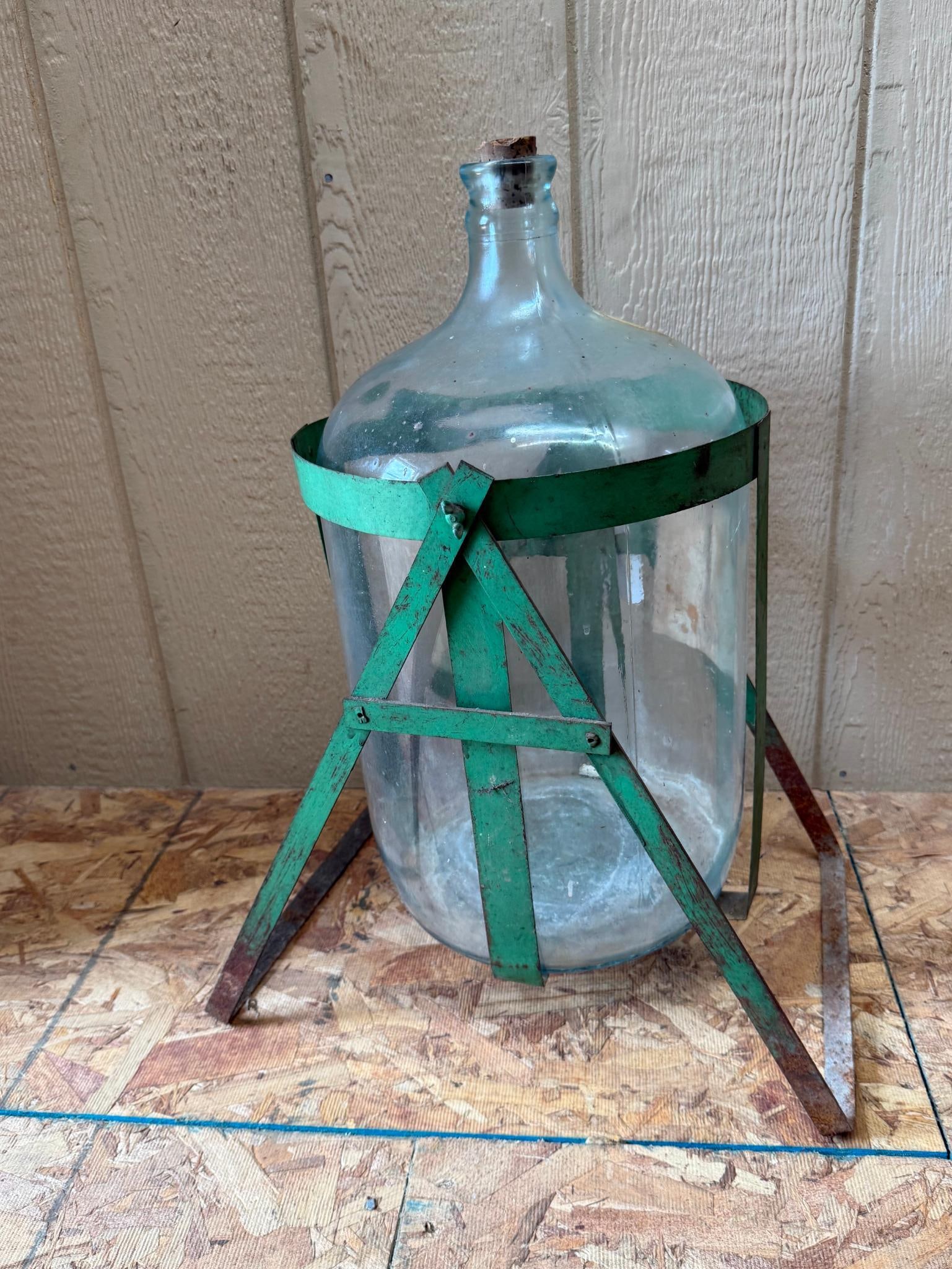 Glass Demijohn in Green Metal Stand: A large clear glass carboy or demijohn secured within a green-painted metal tripod stand. The vessel is made of thick-walled glass with a rounded body and a narrow neck fitted with a weathered and