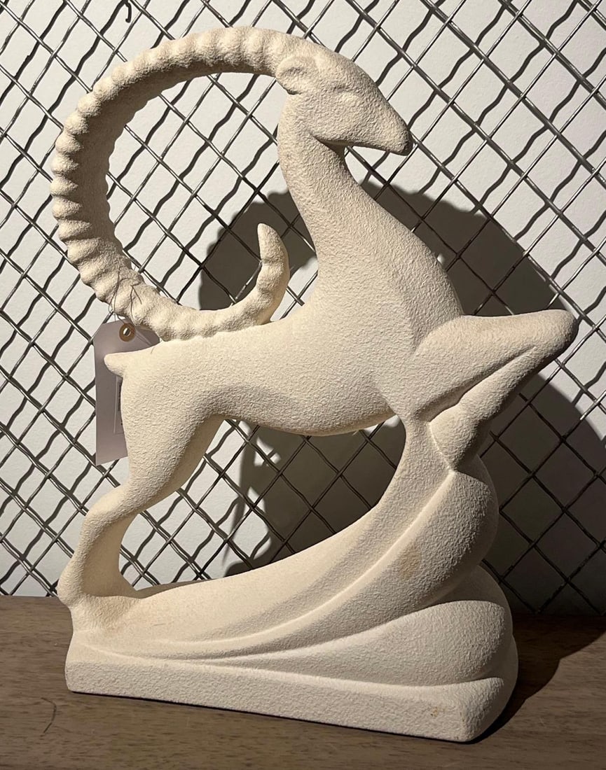 Textured Ceramic Leaping Ibex Sculpture: A stylized ceramic sculpture of a leaping ibex or ram. The figure is depicted in a dynamic, Art Deco-inspired pose with large, curved, ridged horns that form a circular silhouette above its back, moun