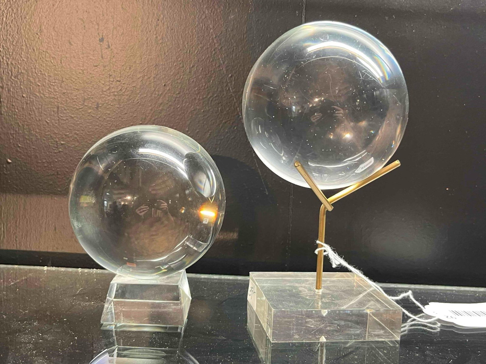 Pair Clear Glass Spheres w/ Stands: A pair of solid, colorless glass spheres, each with a polished surface. One sphere rests on a separate beveled clear glass base. The other sphere is held by a brass-toned metal Y-shaped stand set into
