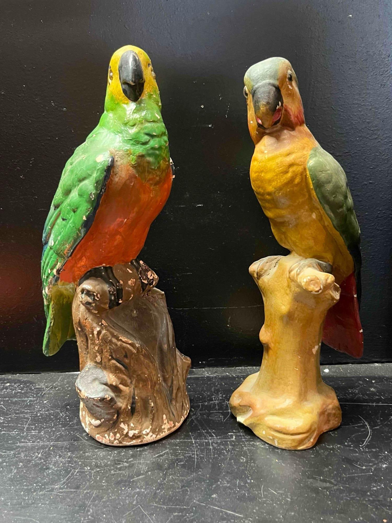 Chalkware Parrot Pair w/ Glass Eyes: A pair of hand-painted chalkware figurines, each depicting a parrot perched atop a textured tree-stump base. The first figure is decorated with a yellow head, green wings, and a bright orange-red brea
