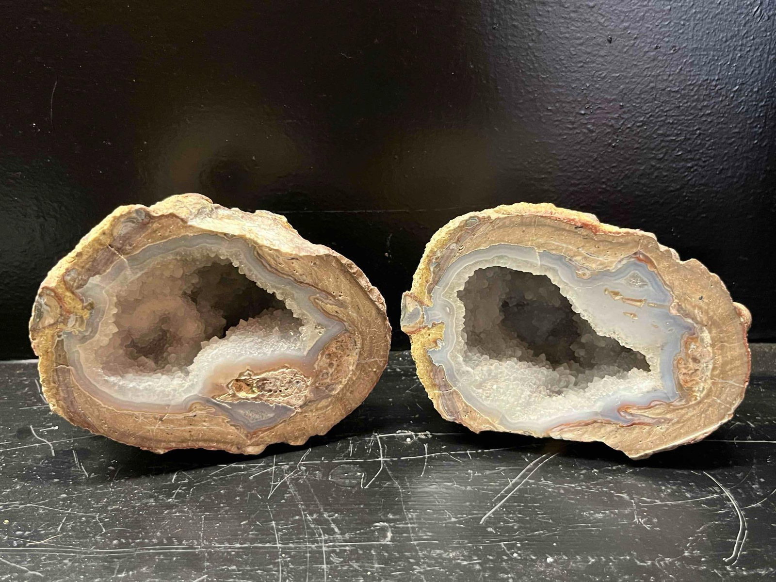Pair Polished Agate & Quartz Geodes: A pair of matched geode halves cut from a single nodule to reveal a complex internal structure. Each specimen features a rugged, natural exterior rind of host rock in shades of buff and reddish-brown.