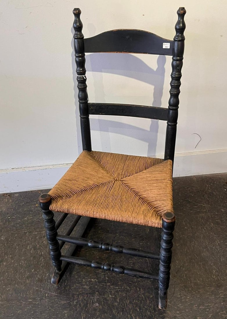Early American Black Ladder Back Rocker: A side rocking chair featuring a black painted wooden frame and a hand-woven rush seat. The backrest is composed of two contoured horizontal slats set between two tall, turned upright posts that termi